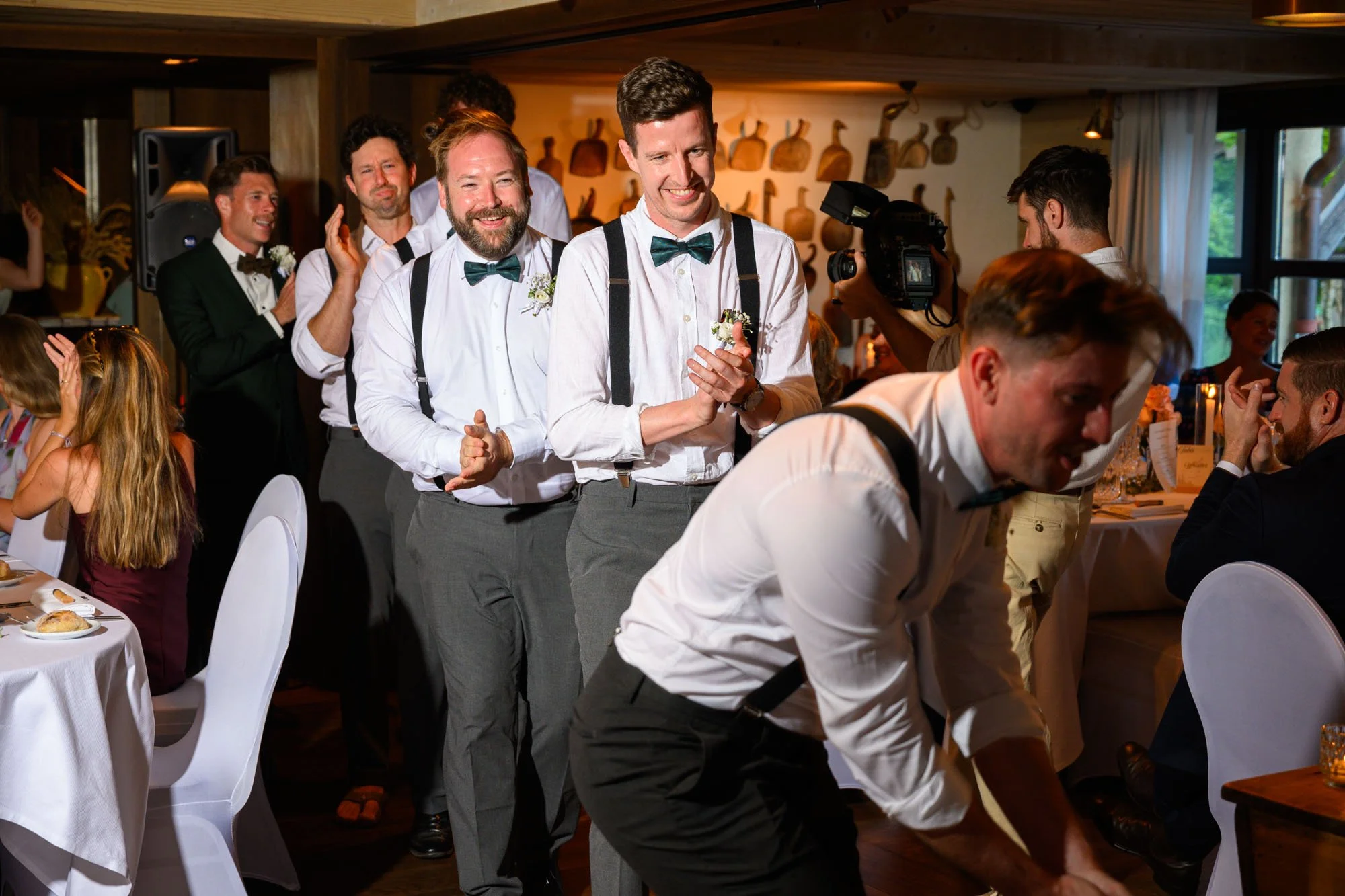 Groupe d'hommes habillés en chemises blanches avec nœuds papillons, applaudissant lors d'un mariage dans une salle décorée avec des œuvres en bois au mur.