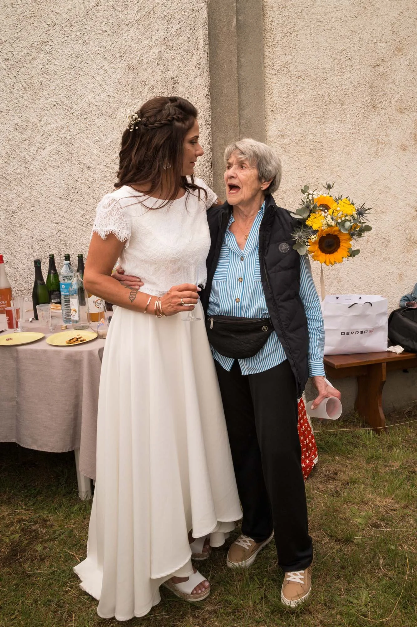 Une jeune femme en robe blanche et une femme âgée en conversation lors d'un événement en plein air, avec une table de boissons derrière elles.