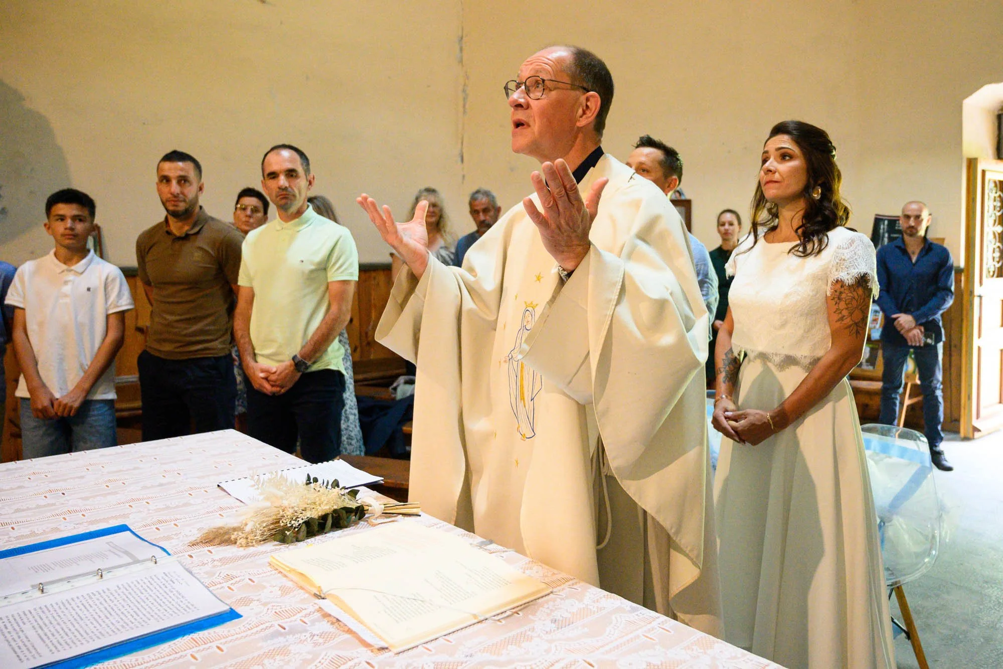 Un prêtre devant un groupe de personnes lors d'une cérémonie religieuse, avec un livre ouvert et des fleurs sur la table.