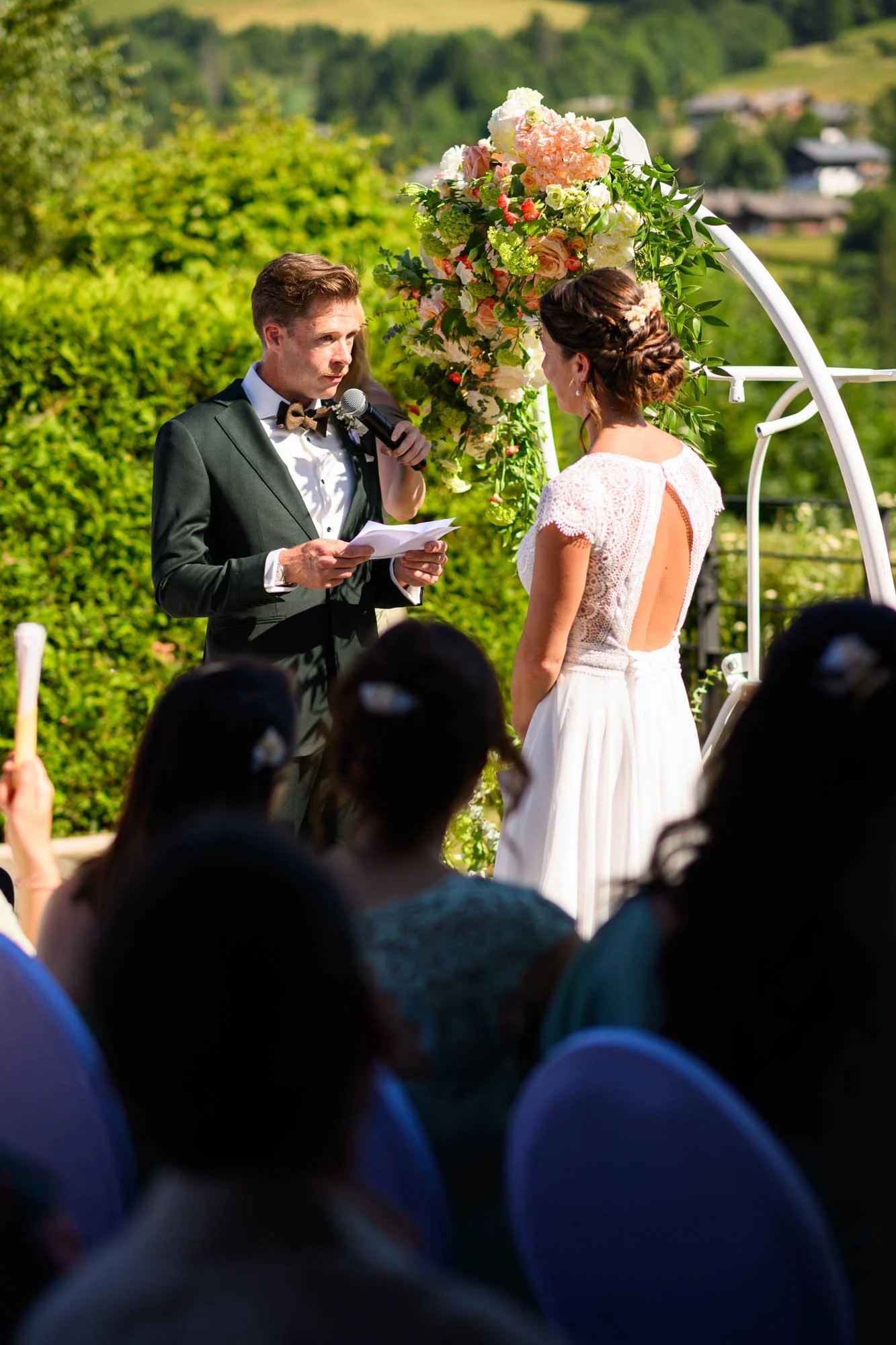 Un couple échange leurs vœux de mariage lors d'une cérémonie en extérieur, avec un officiant lisant un discours, entouré de fleurs et de paysage vert.