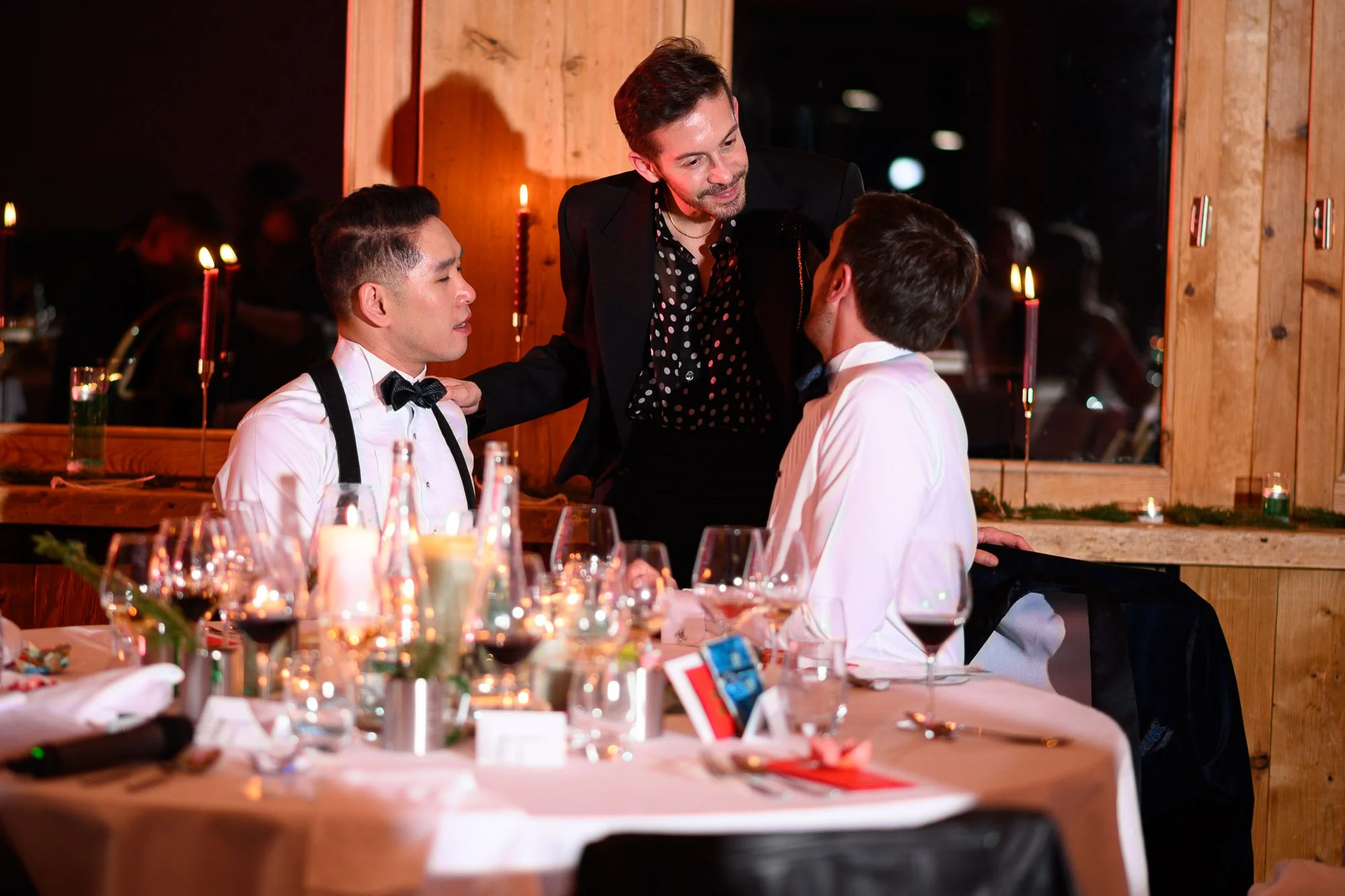 Deux hommes en smoking discutent avec une autre homme qui les regarde avec un sourire, lors d'un dîner dans un restaurant chic, avec des bougies sur la table.
