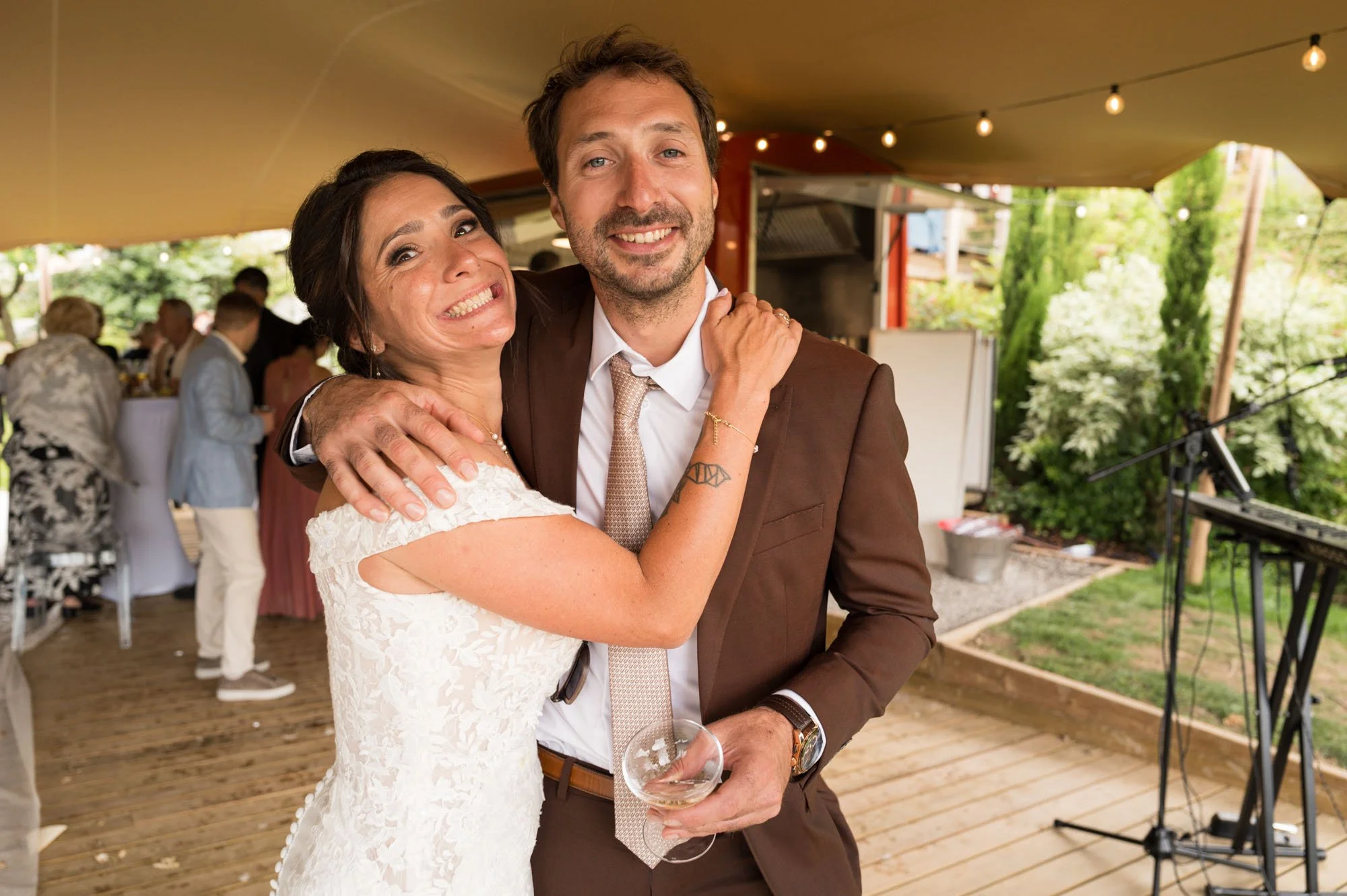 Un couple souriant lors d'une fête ou d'un mariage, en train de s'embrasser et de poser pour la photo dans un espace extérieur.