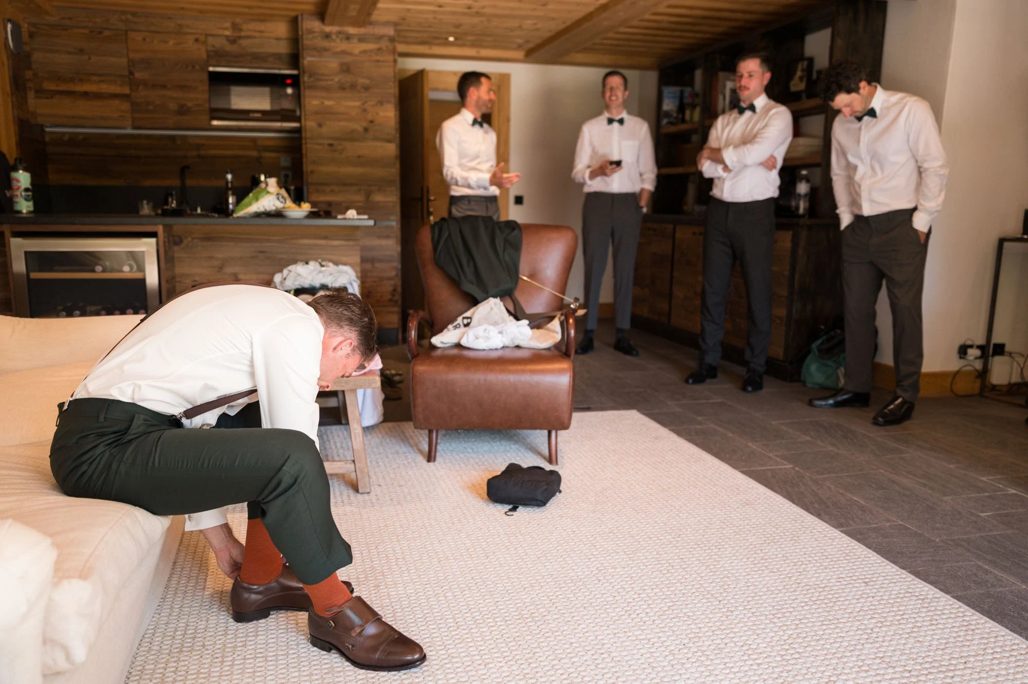 Homme en costume blanc et chaussures marron, assis et en train de s'ajuster ses chaussures, dans une pièce à l'intérieur d'une maison, avec quatre autres hommes en costume blanc avec nœud papillon, debout à l'arrière, certains utilisant leur téléphon