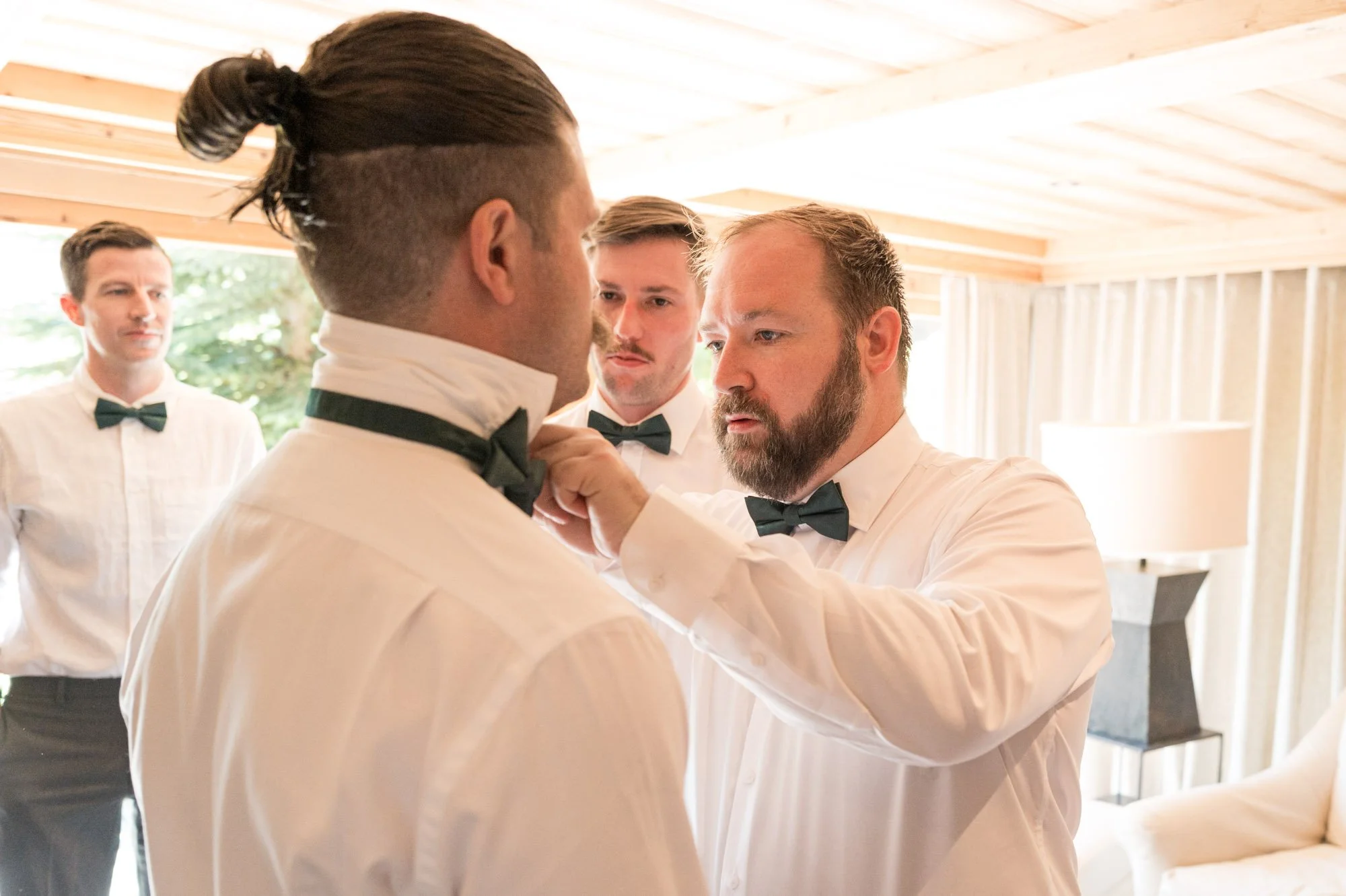 Homme ajustant le nœud papillon d'un autre homme dans une pièce lumineuse, avec deux autres hommes en arrière-plan, tous portant des chemises blanches et des nœuds papillons noirs.