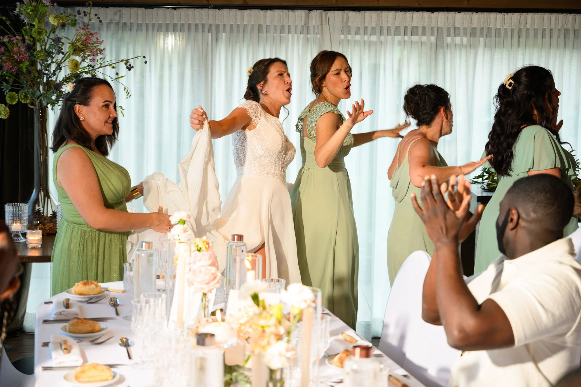 Fives women, some in party dresses, singing or speaking at a wedding reception, with a man clapping at the table in the foreground.