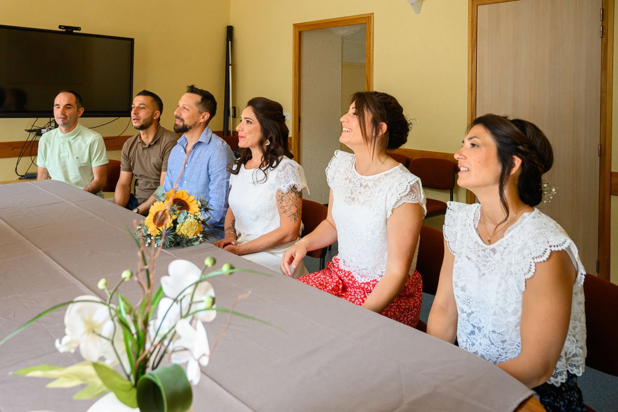 Groupe de personnes assises autour d'une table lors d'une réunion ou d'un événement, avec un vase de fleurs en premier plan.