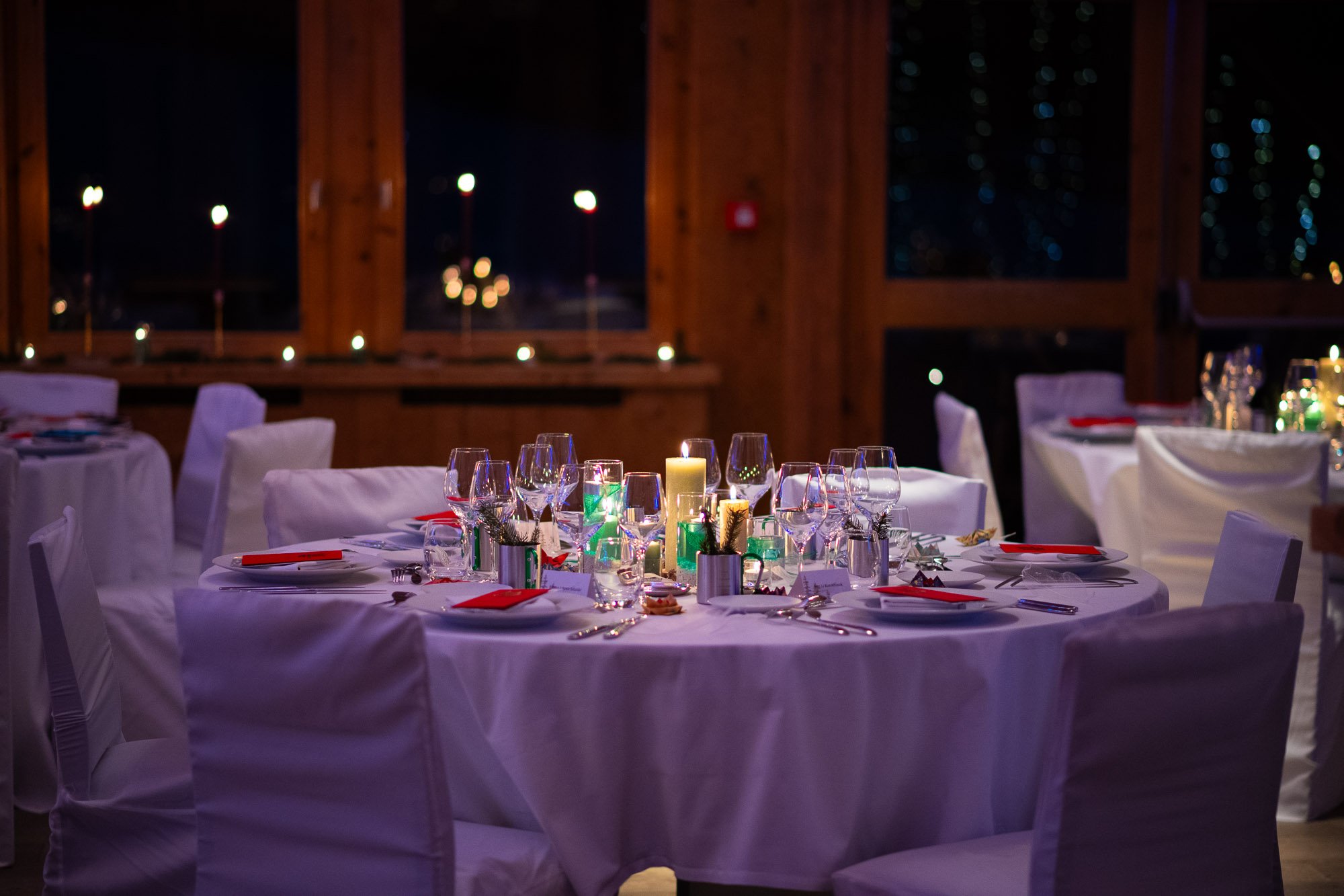 Table de fête élégamment dressée avec des bougies et des verres, ambiance chaleureuse et festive, fenêtres en arrière-plan avec vues nocturnes.
