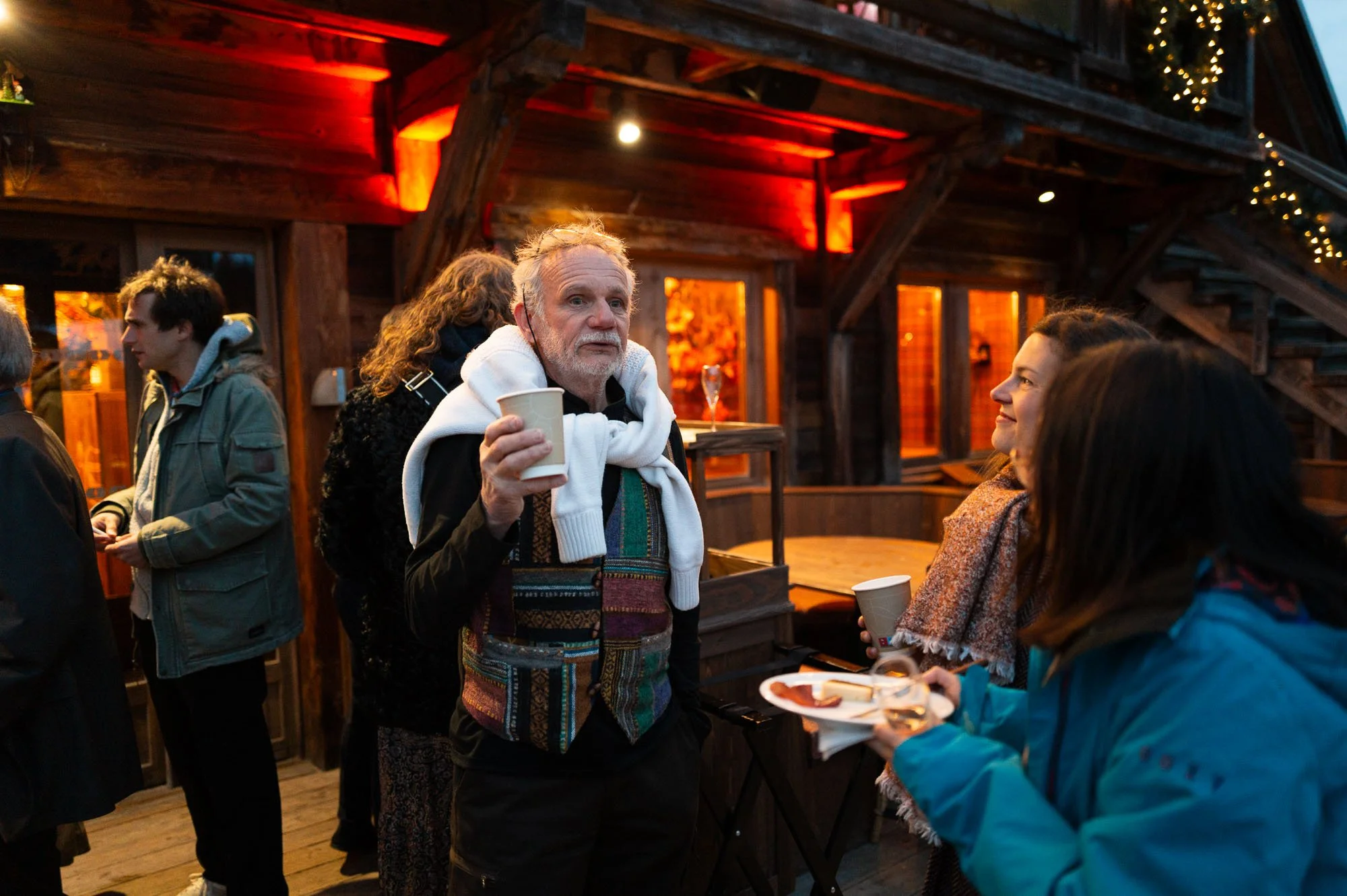 Groupe de personnes profitant d'une fête en extérieur avec une ambiance chaleureuse et décorations lumineuses dans une maison en bois.