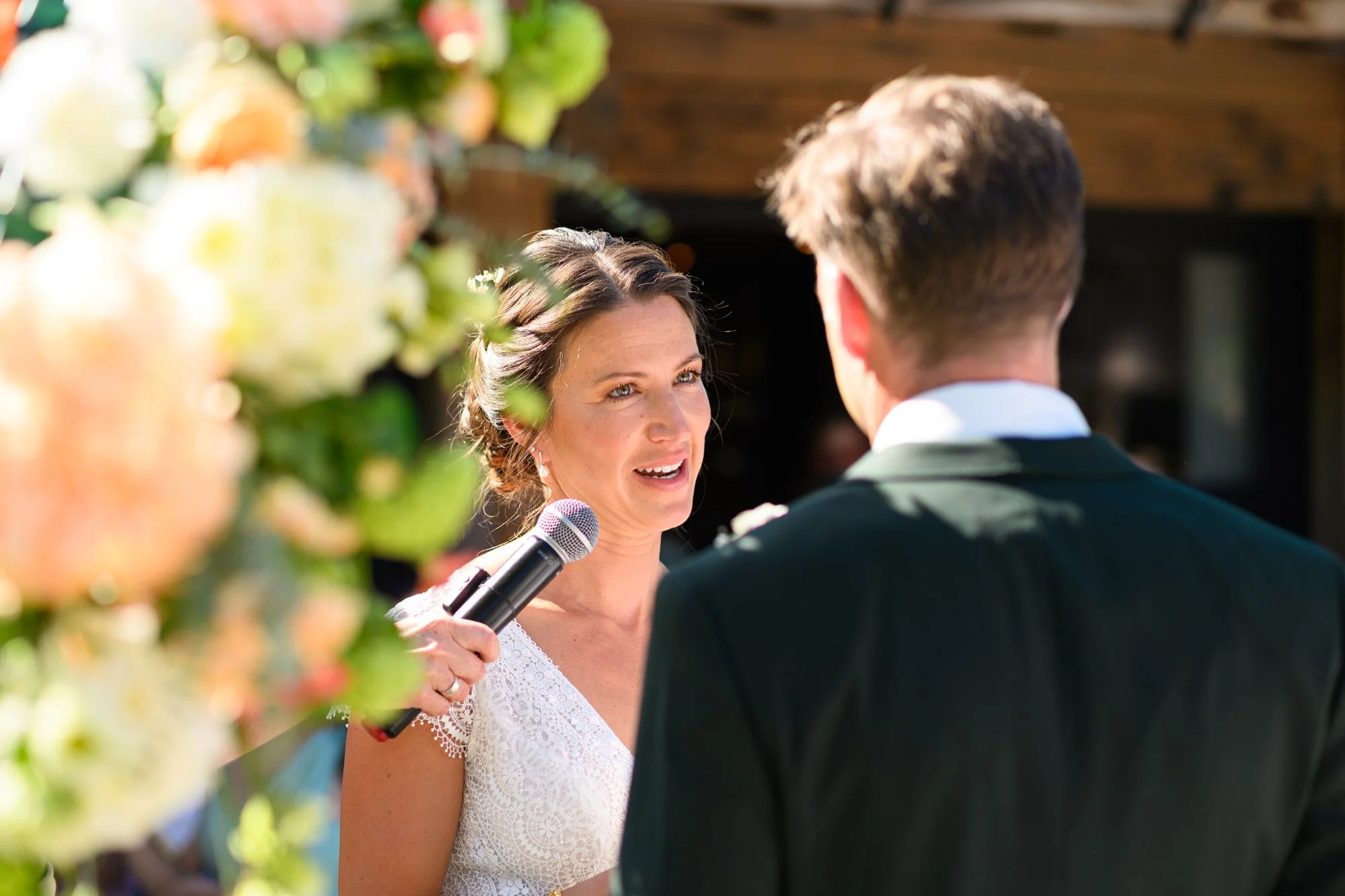 Une femme avec un microphone, en robe blanche, regardant un homme en costume lors d'un mariage en plein air.