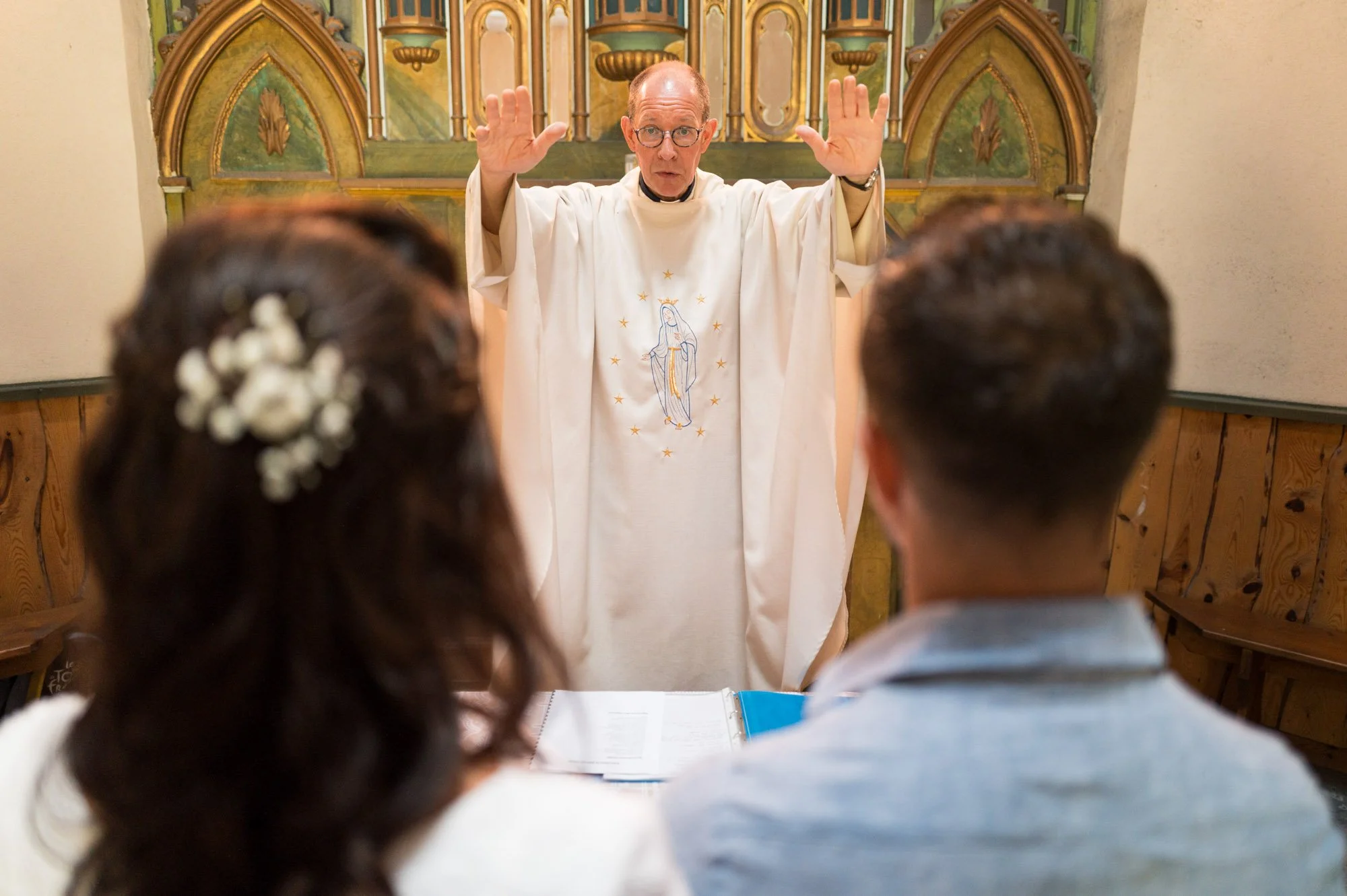 Un prêtre célèbre un mariage religieux avec un couple, la femme a des fleurs blanches dans les cheveux.