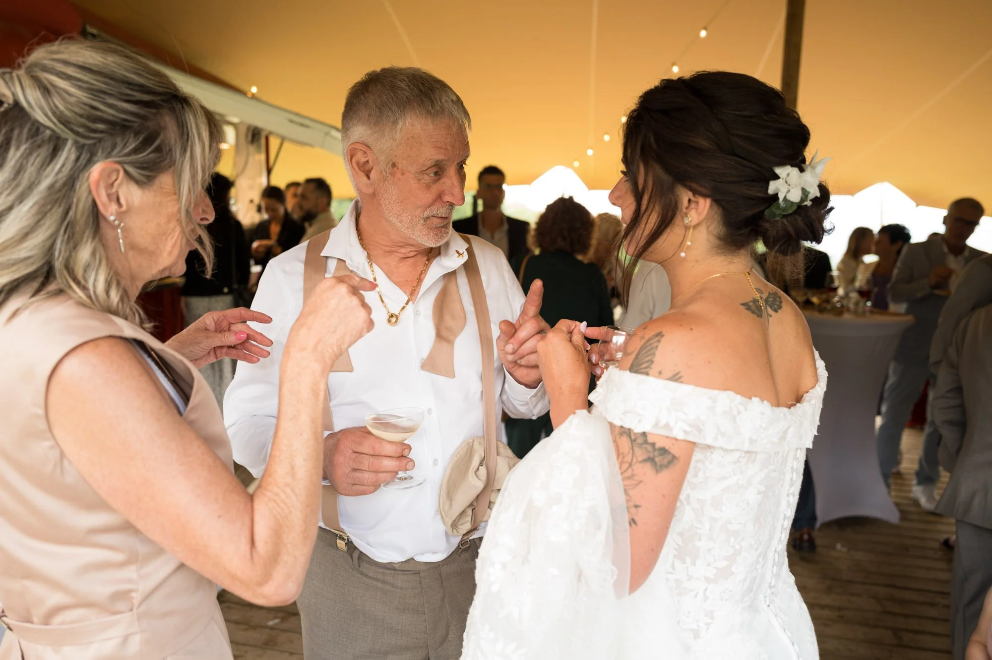 Des invités discutent lors d'un mariage sous une tente, avec une mariée en robe blanche et des fleurs dans ses cheveux.