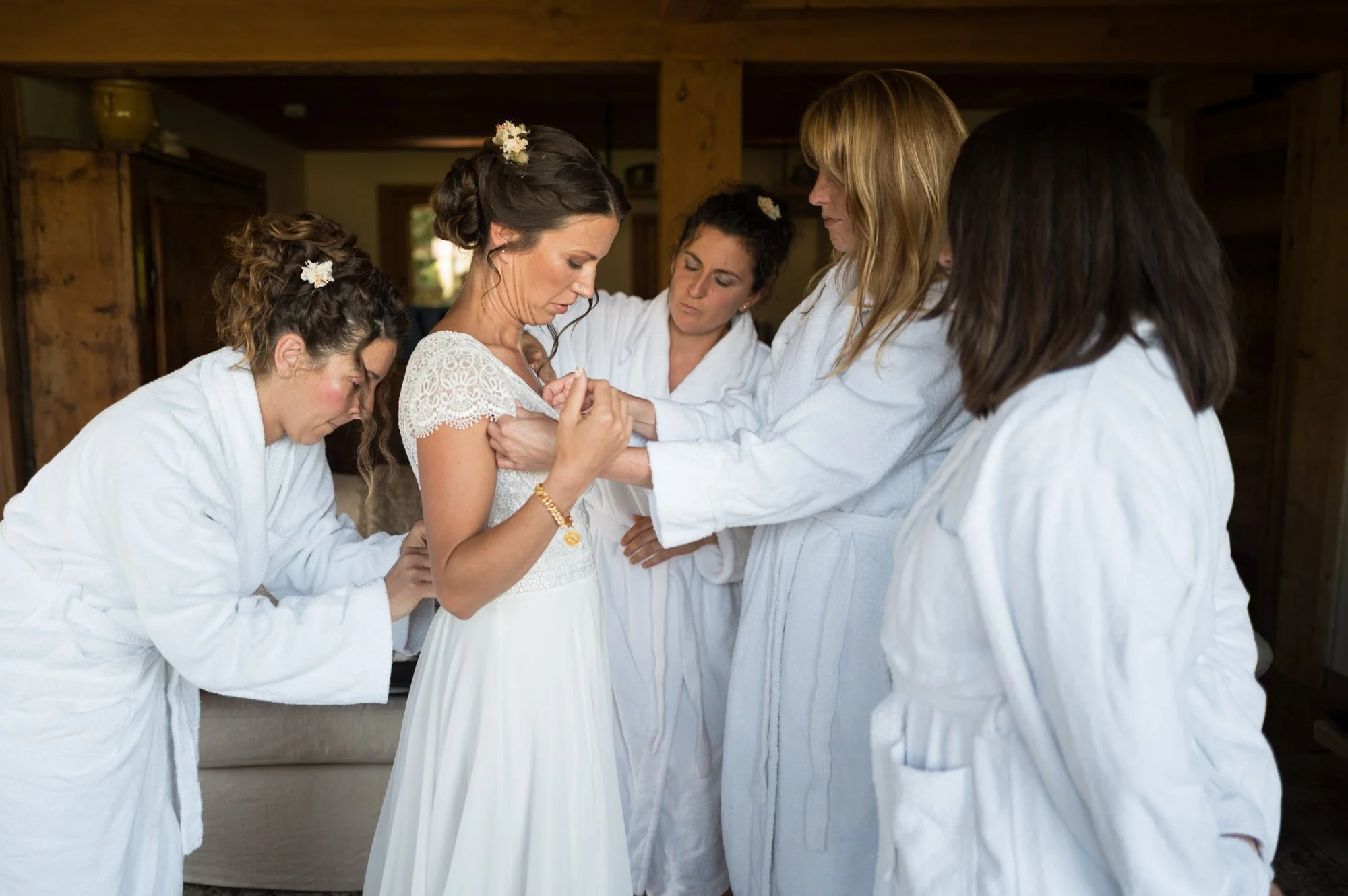 Femme en robe de mariée entourée de demoiselles d'honneur en peignoirs blancs, aidant à préparer la mariée.