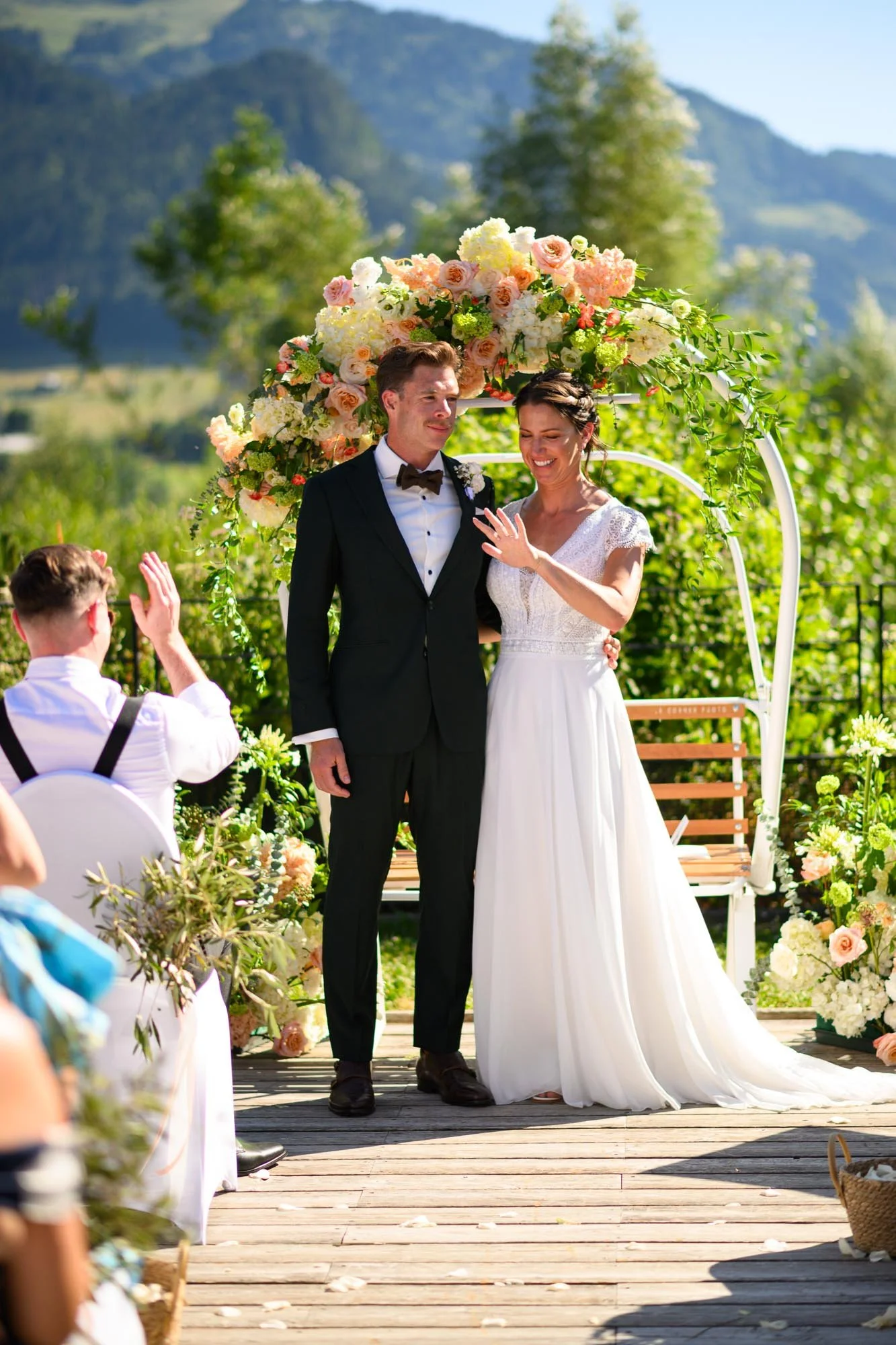 Un mariage en plein air avec le couple marié sous une arche fleurie, la mariée en robe blanche et le marié en costume noir, entourés de décorations florales et d'un paysage naturel en arrière-plan.