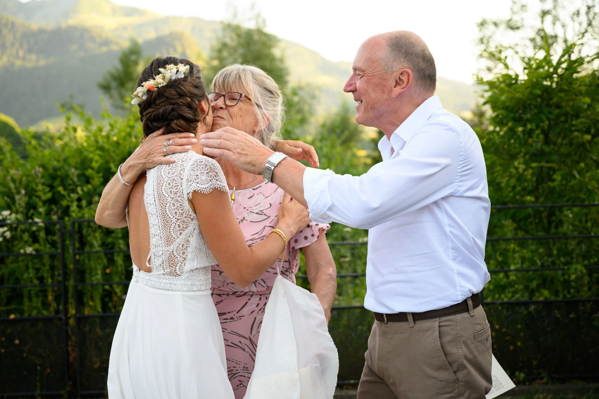 Une jeune femme en robe de mariée embrasse un vieil homme et une femme plus âgée lors d'une cérémonie extérieure, dans un endroit verdoyant avec des montagnes en arrière-plan.