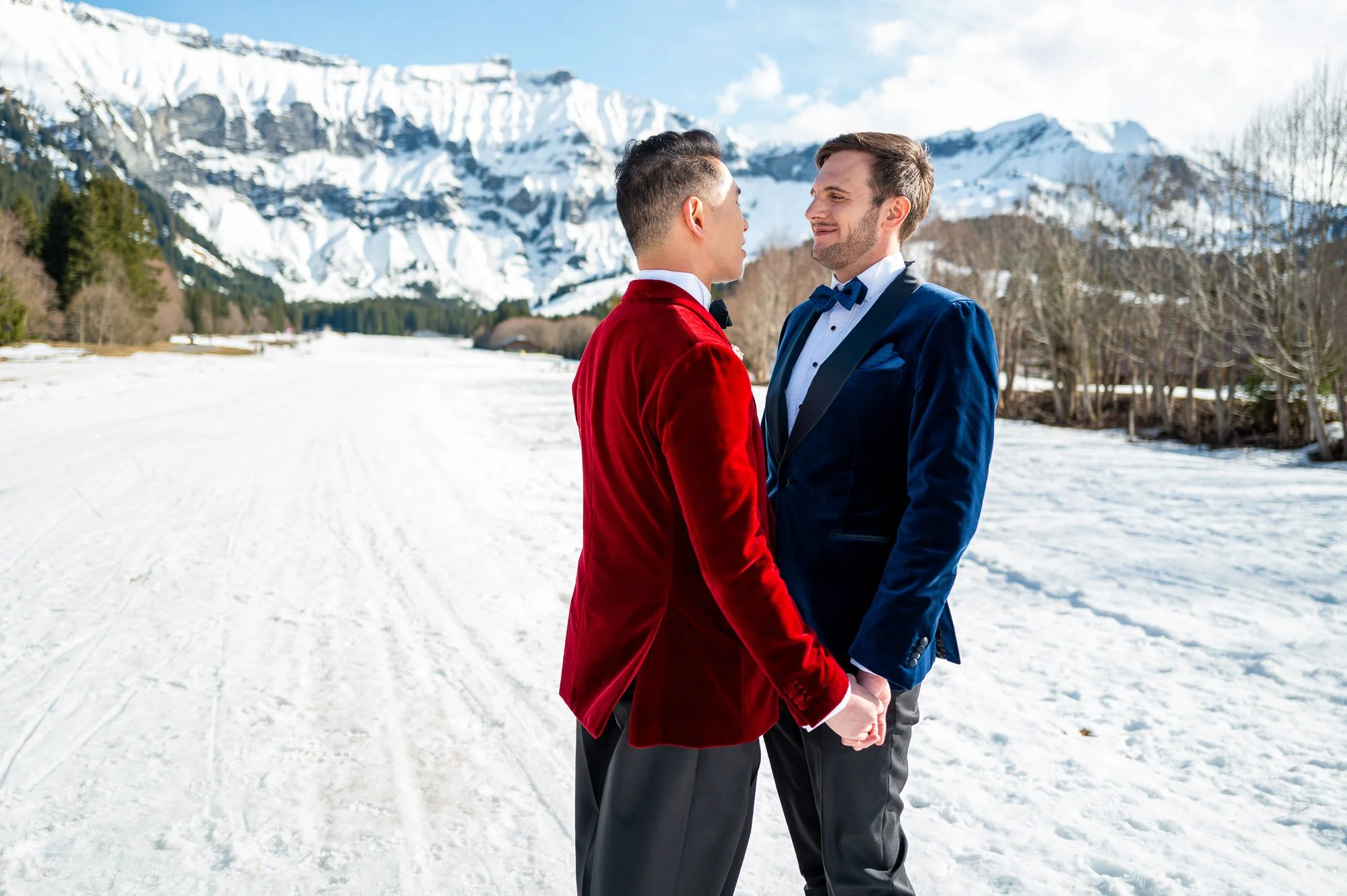 Deux hommes habillés en smoking, tenant la main, face à face dans un paysage enneigé avec des montagnes en arrière-plan.