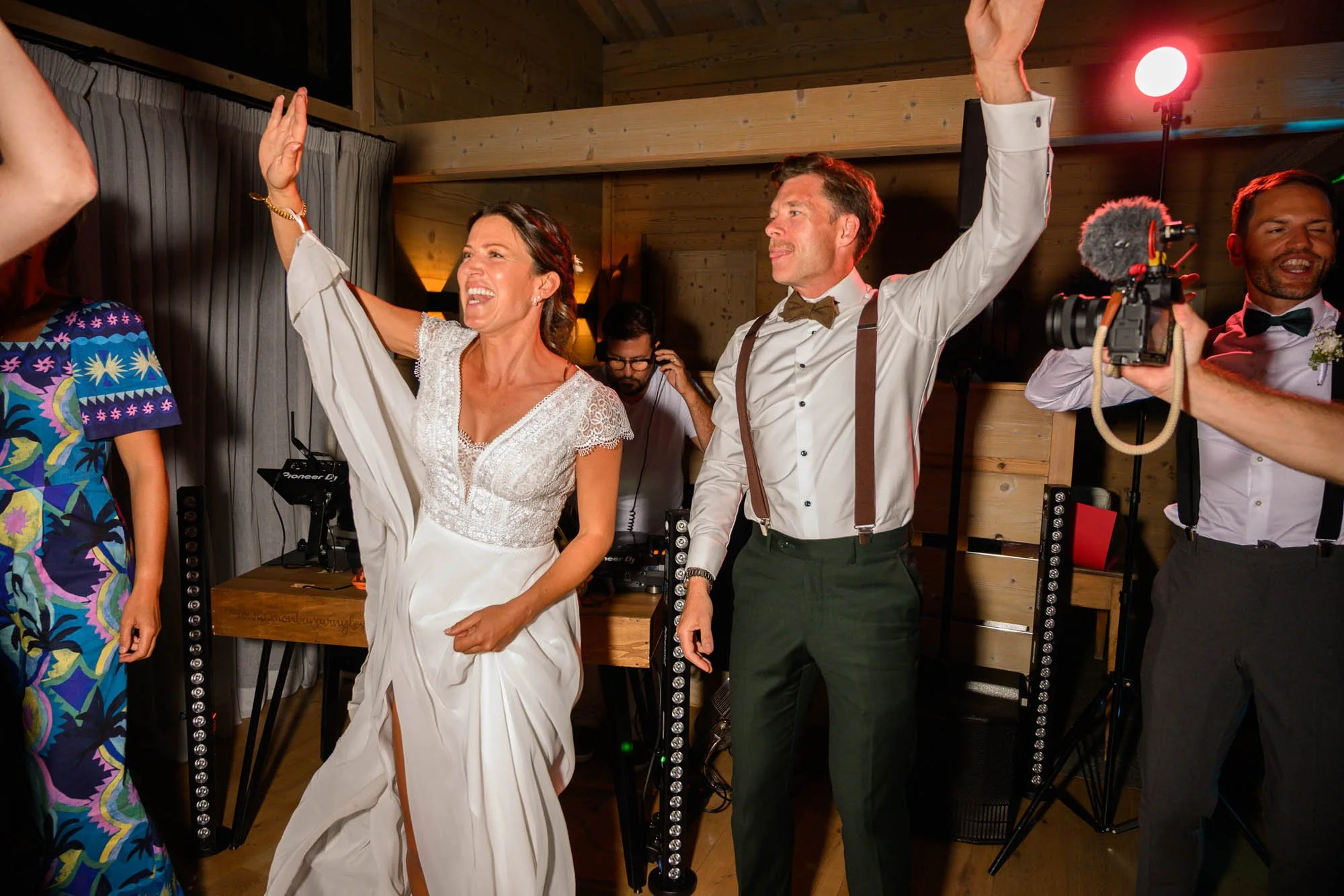 Un couple en costumes de mariage danse et chante lors de leur réception, entourés d'amis et de musiciens dans une salle en bois.