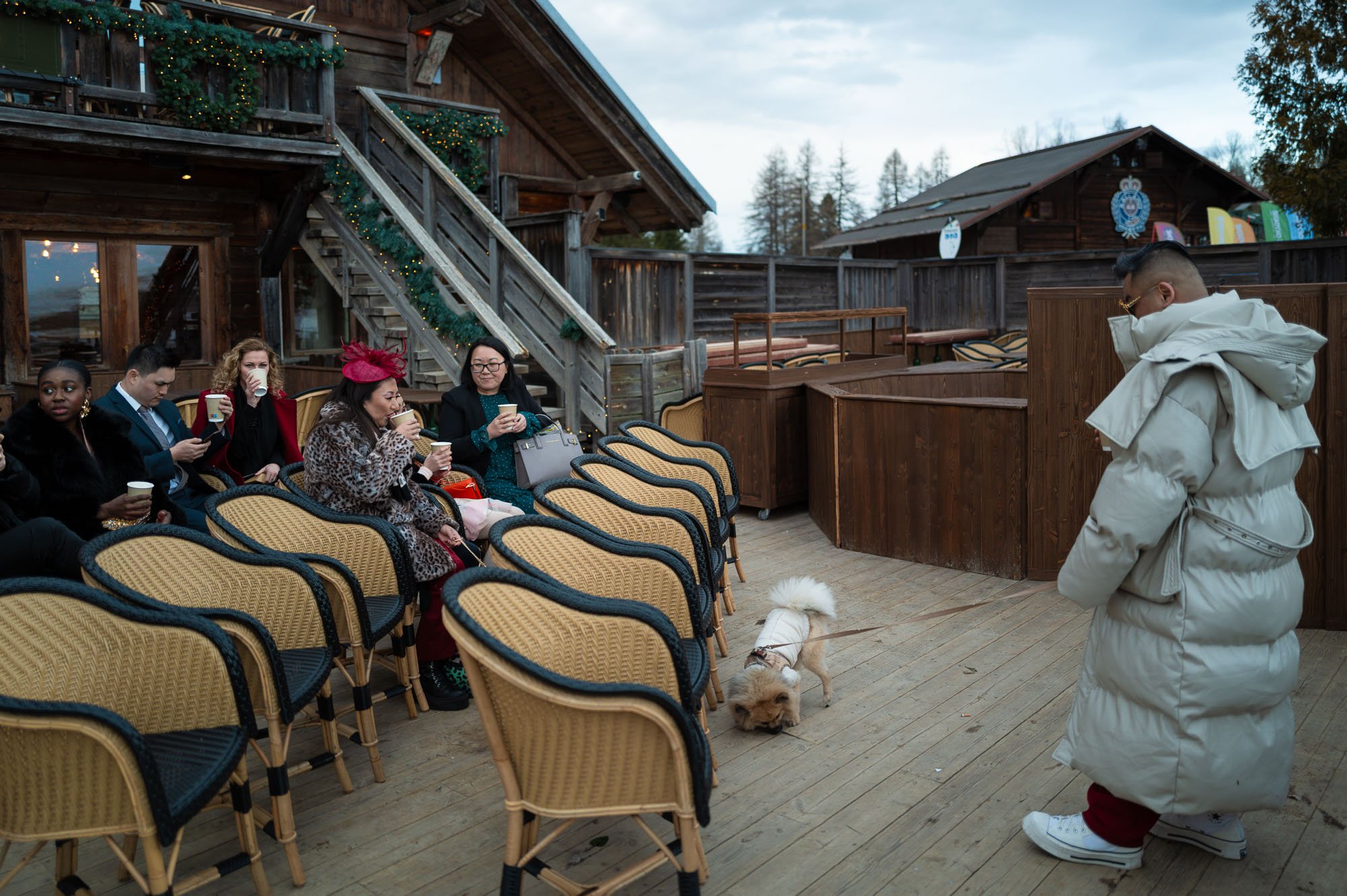 Groupe de personnes assises en extérieur, dégustant des boissons, avec un chien et un homme debout portant un manteau épais.