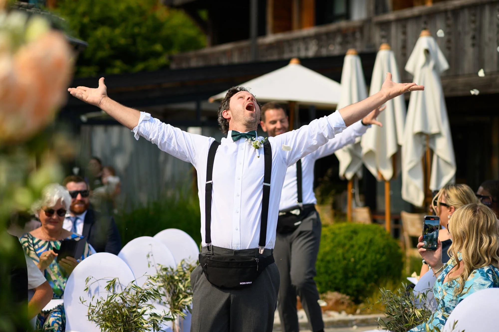 Un homme en costume blanc avec un nœud papillon vert, levant les bras en signe de victoire lors d'un mariage en plein air, avec des invités qui le photographient.
