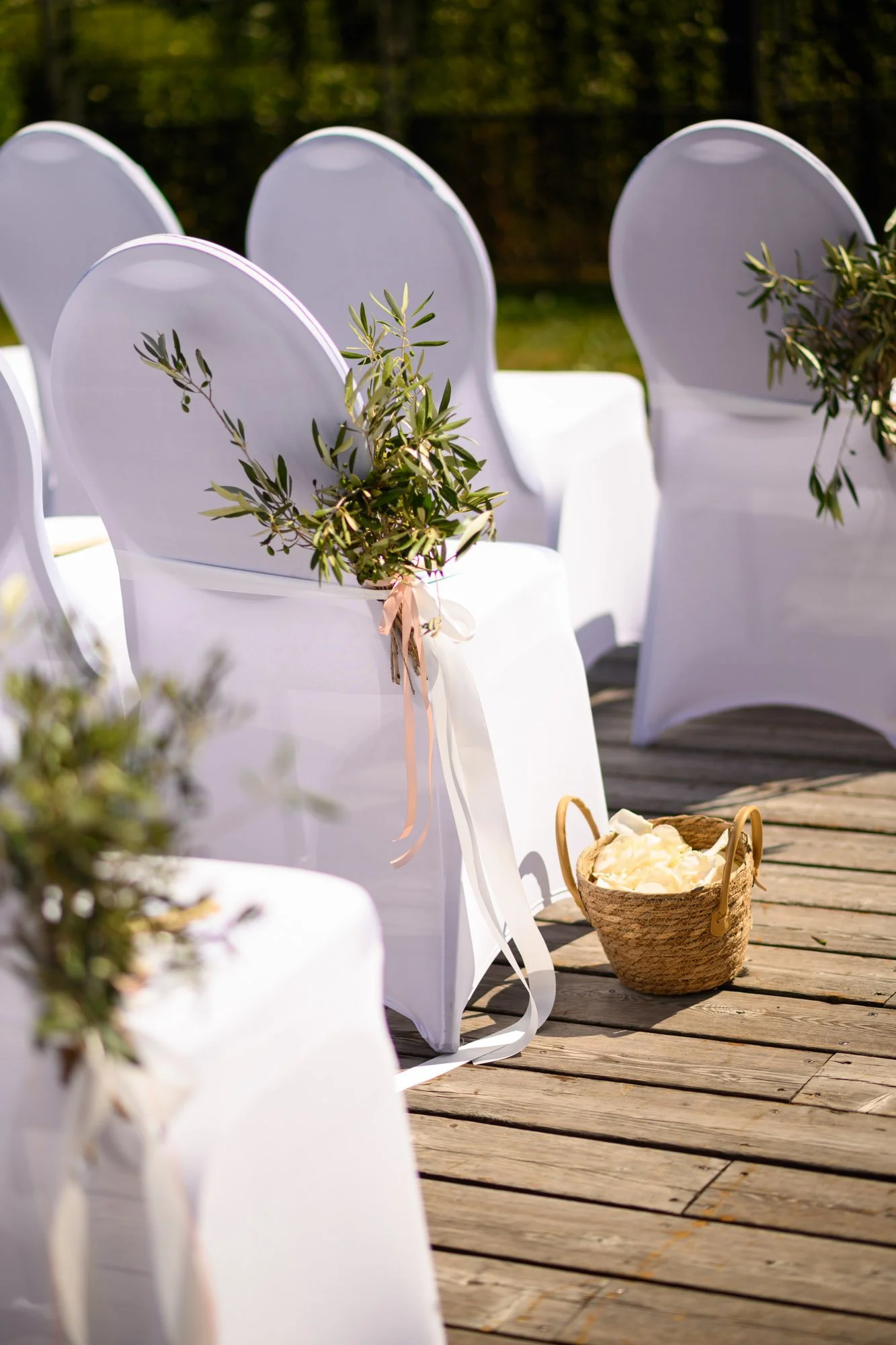 Chaises blanches décorées avec des branches d'olivier et des rubans, disposées sur une terrasse en bois, probablement pour une cérémonie de mariage.
