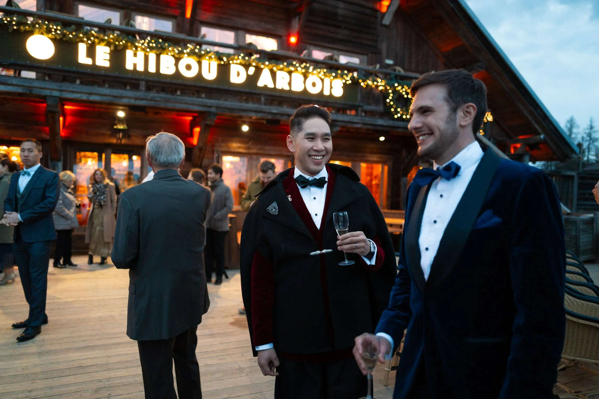 Groupe de personnes en tenue élégante lors d'une soirée extérieure devant un bâtiment en bois avec un panneau lumineux disant "Le Hibou d'Arbois". Deux hommes en costume discutent, tenant des verres de champagne, souriants.