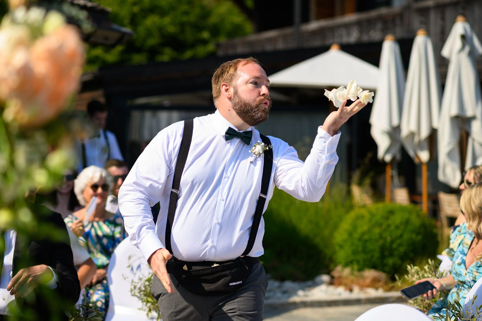 Un homme avec une chemise blanche, un nœud papillon noir, et des bretelles, souffle des fleurs lors d'un mariage en plein air, entouré de invités assis.