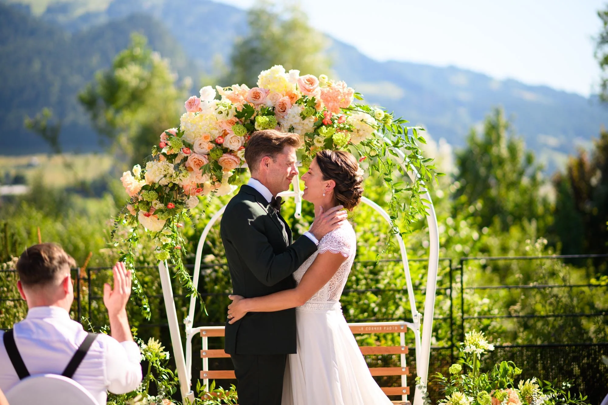 Un couple de mariés s'embrasse lors de la cérémonie en plein air, entouré d'une arche de fleurs colorées, avec deux hommes en arrière-plan applaudissant.