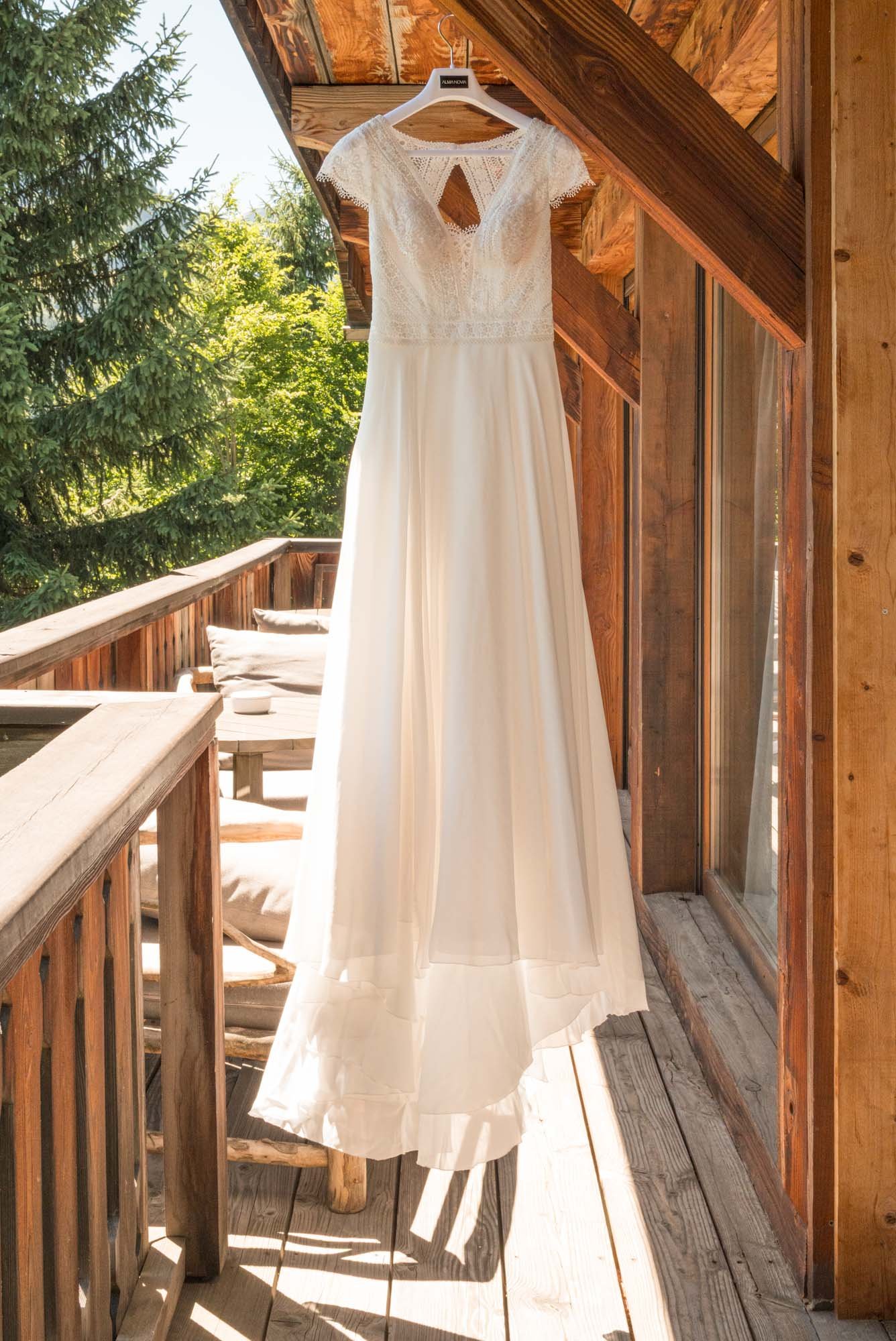 Robe de mariage blanche suspendue à un crochet sur un balcon en bois, avec un paysage de forêt en arrière-plan.