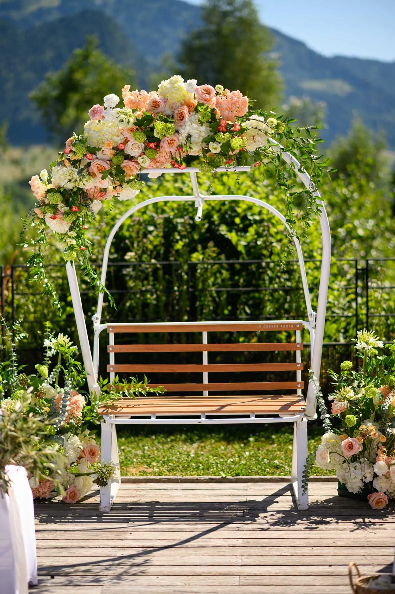 Baldaquin de mariage en métal blanc décoré de fleurs pastel, installé sur une terrasse en bois avec paysage naturel en arrière-plan.