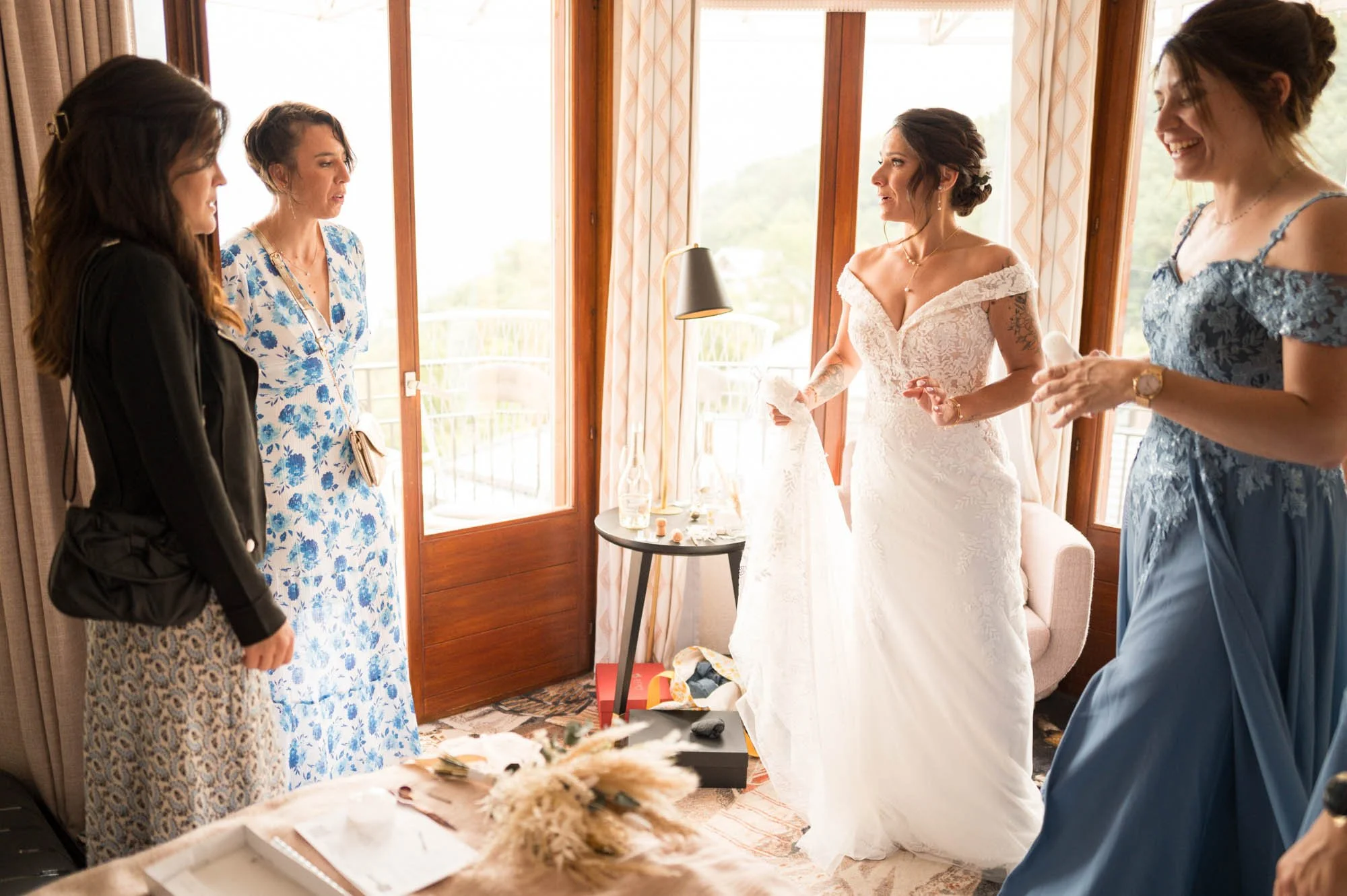 Cinq femmes dans une chambre lumineuse préparant une mariée en robe blanche avec des détails en dentelle. La mariée se tient au centre, entourée de ses amies, toutes souriantes et habillées élégamment.