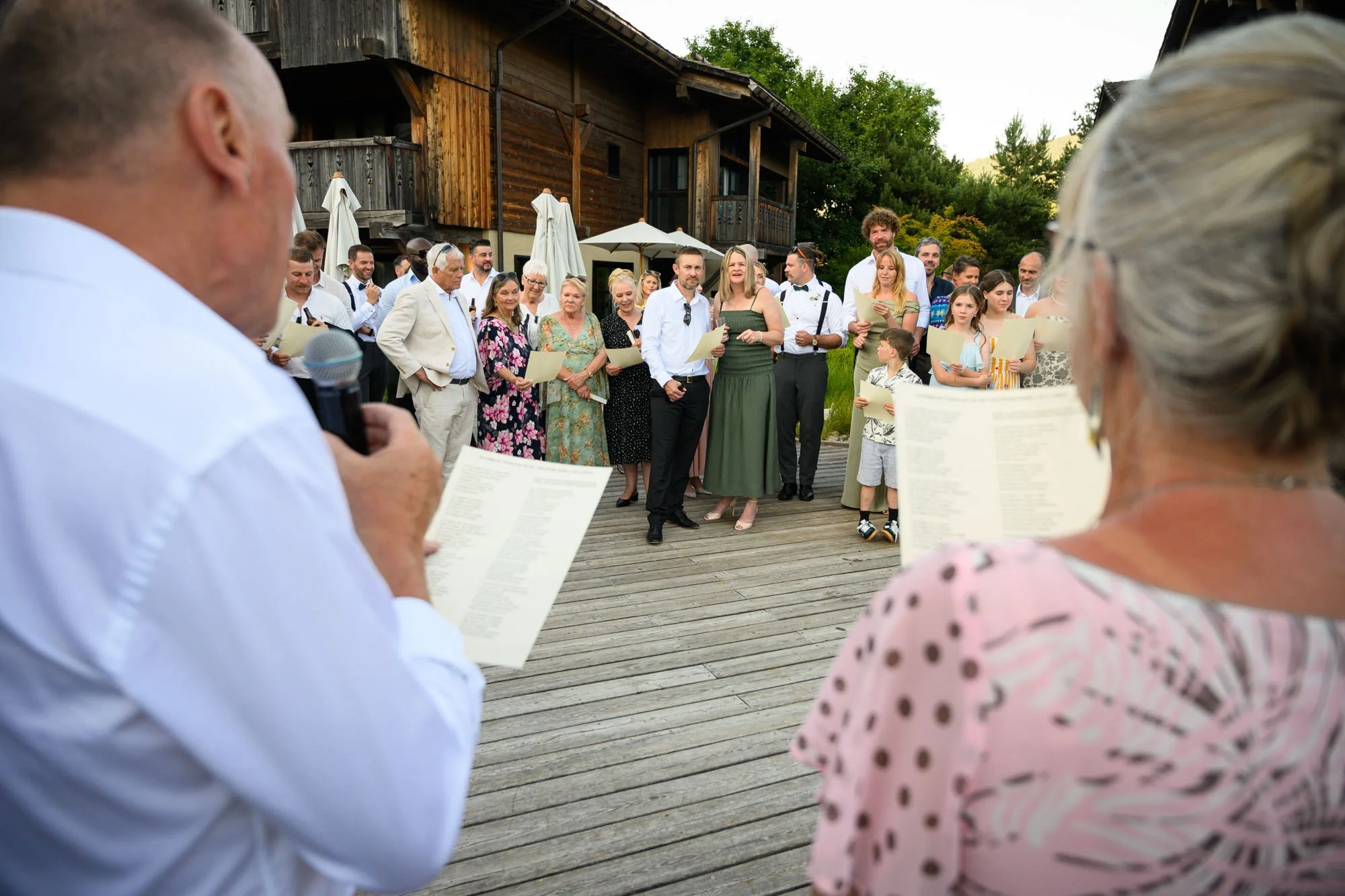 Groupe de personnes lors d'une cérémonie en extérieur, certains lisant des discours, entourés de maisons en bois et de végétation.