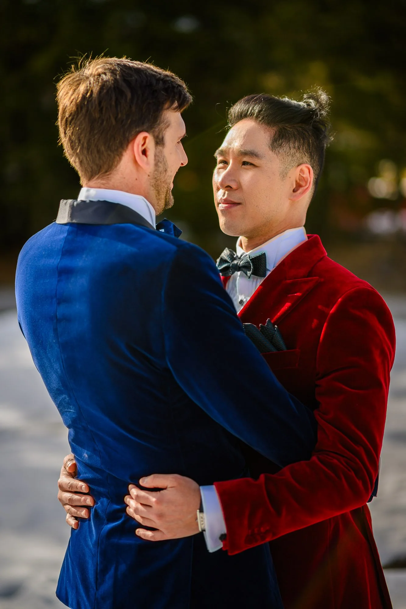 Deux hommes en costumes formels, un en costume bleu et l'autre en costume rouge, se regardent tendrement lors d'une rencontre en plein air, probablement pour une occasion spéciale ou romantique.