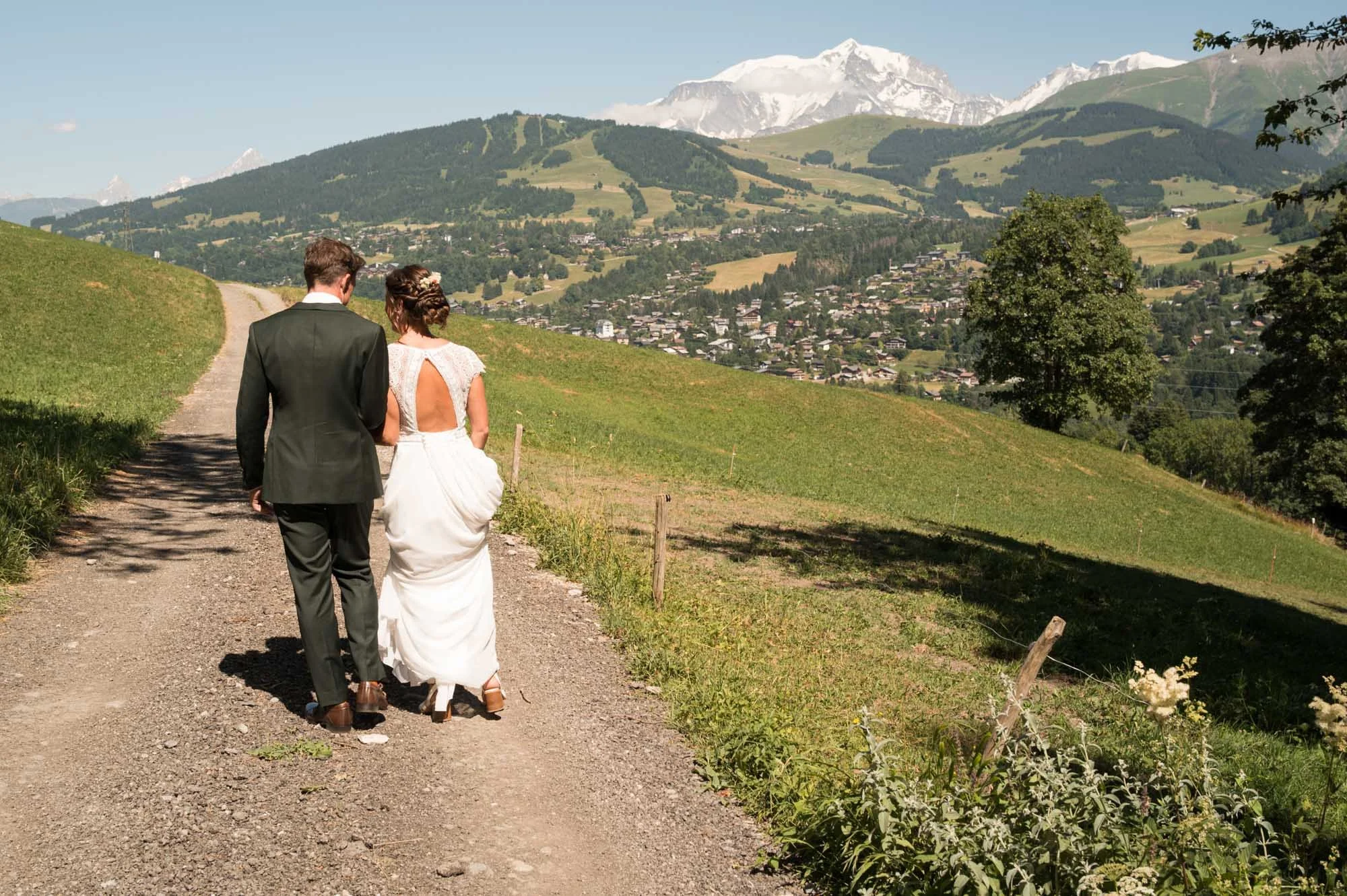 Un couple vêtu d'un costume de marié marche sur un chemin de terre dans une campagne vallonnée avec des montagnes enneigées en arrière-plan.