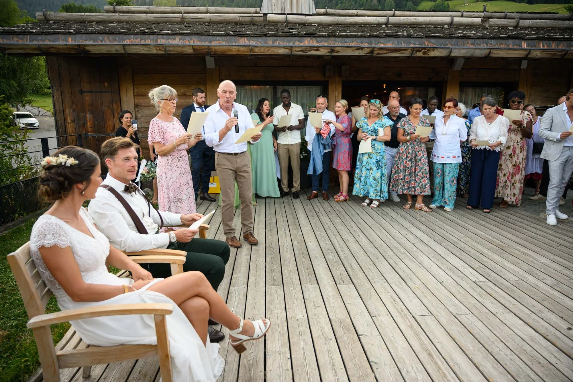 Un groupe de personnes participe à une cérémonie de mariage en extérieur, avec deux personnes assises, probablement le marié et la mariée, et plusieurs autres personnes se tenant derrière eux, en train de lire ou d'écouter.
