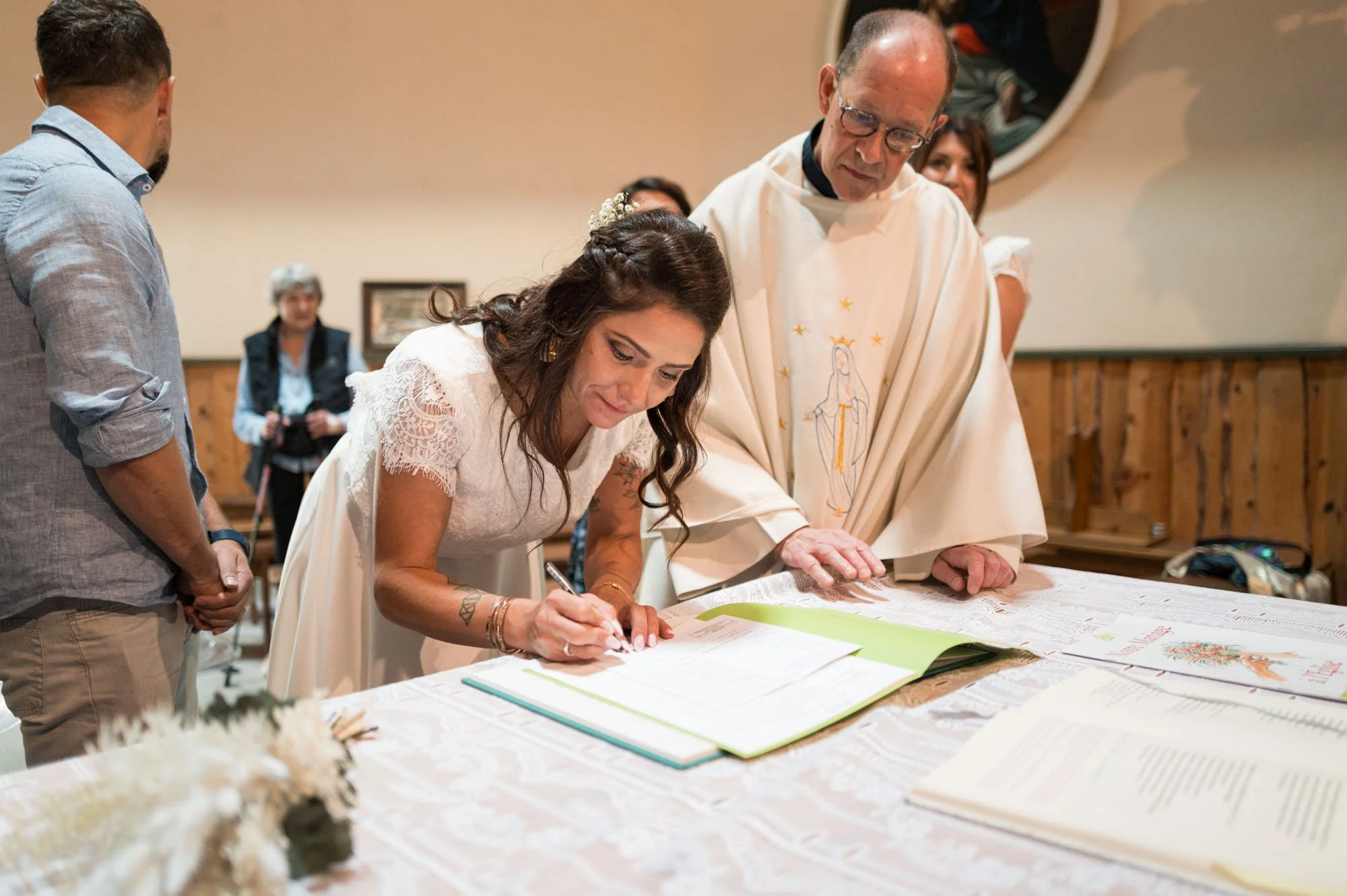 Une femme signe un registre lors d'une cérémonie religieuse, avec un prêtre ou un célébrant à ses côtés, en présence de plusieurs personnes. La scène se déroule dans une salle avec des murs en bois.