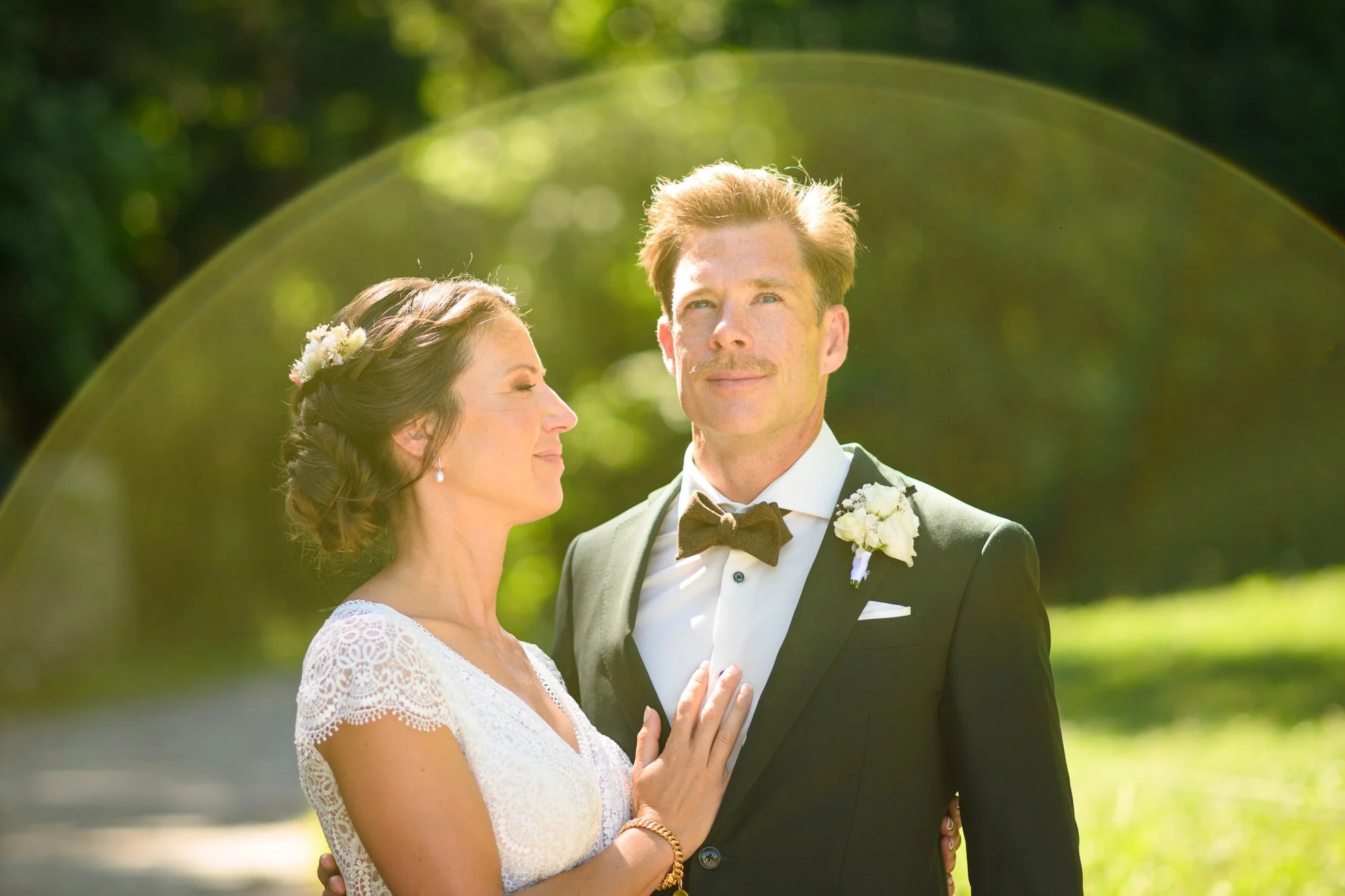 Un couple de mariés posant dehors lors de leur mariage. La femme porte une robe blanche en dentelle et a les yeux fermés, le marié porte un costume noir avec un nœud papillon et une petite boutonnière blanche. En arrière-plan, il y a de la verdure et