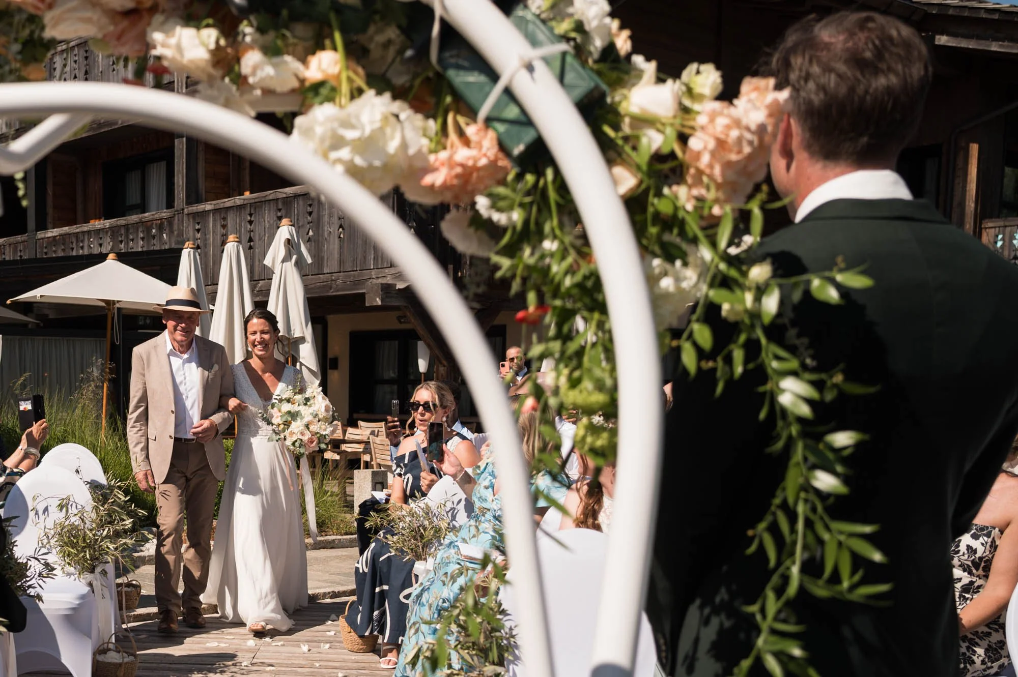 Cérémonie de mariage en plein air avec le couple marié marchant sous un arc fleuri, entouré d'invités assis et prenant des photos, décorations florales, parasols et bâtiment en bois en arrière-plan.