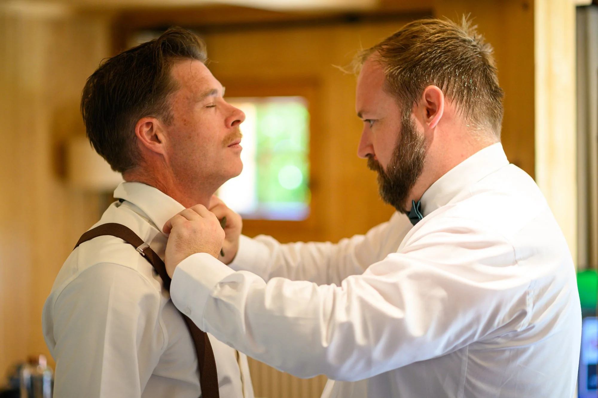 Un homme en costume blanc ajustant la cravate d'un autre homme, également en costume blanc, dans une pièce avec des murs en bois.