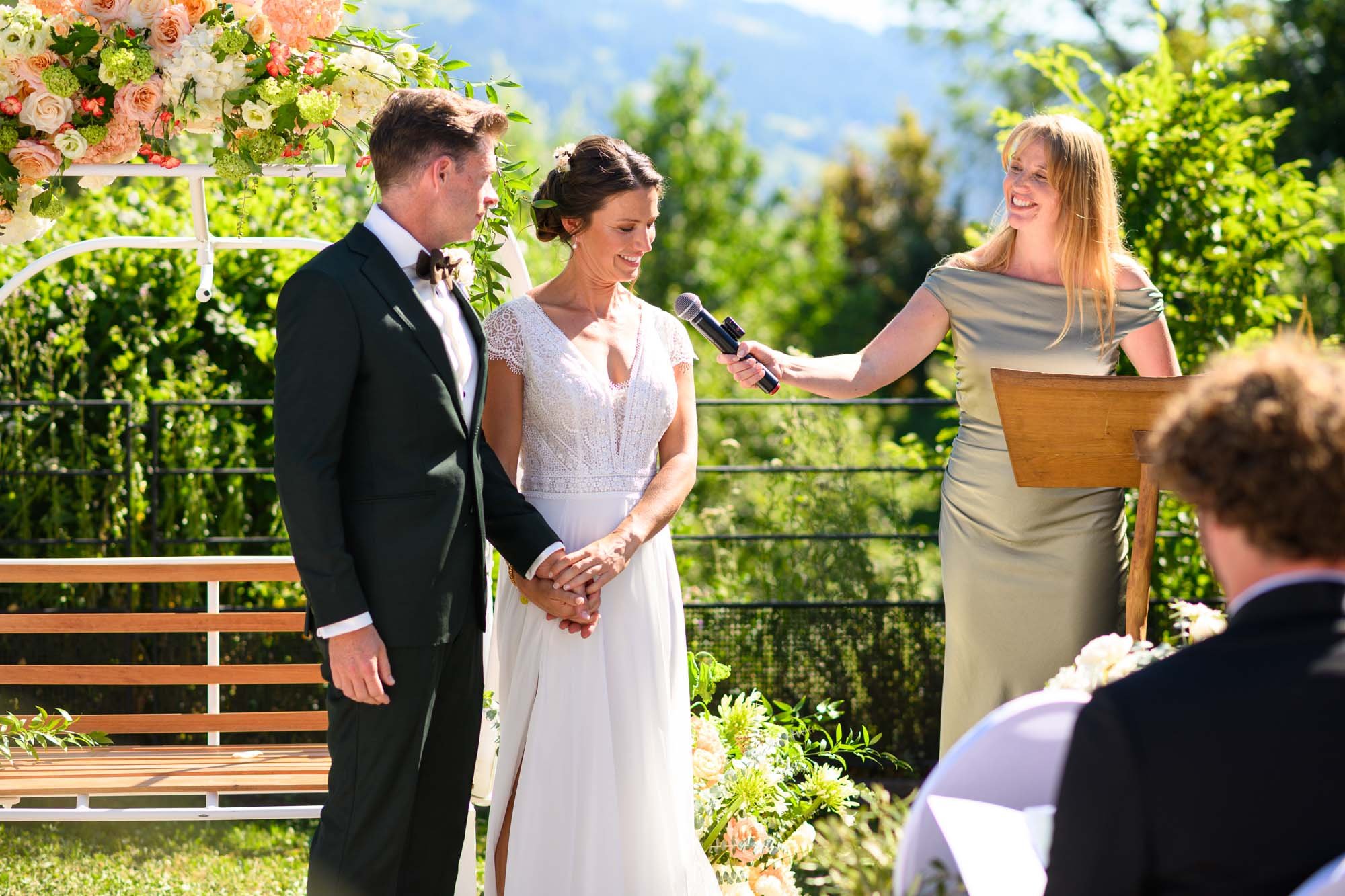 Un couple en costume de mariage tenant la main lors d'une cérémonie en plein air, devant un officiant féminin qui tient un micro, avec une décoration florale devant eux, sous un ciel bleu et entouré de verdure.