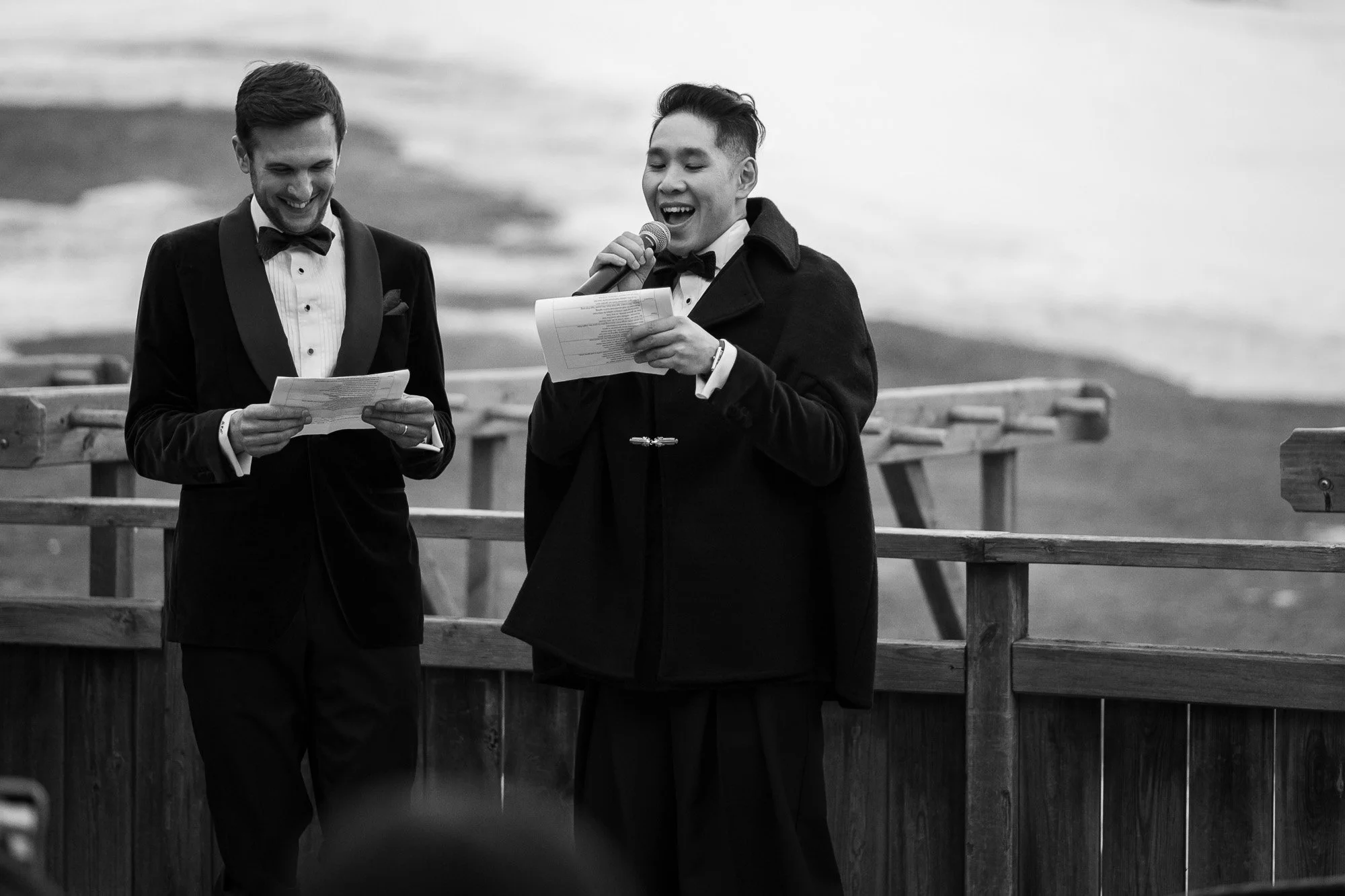 Deux hommes en costume et manteau noir, souriants, parlant dans un micro et tenant des papiers, sur un quai en bois près de la mer.