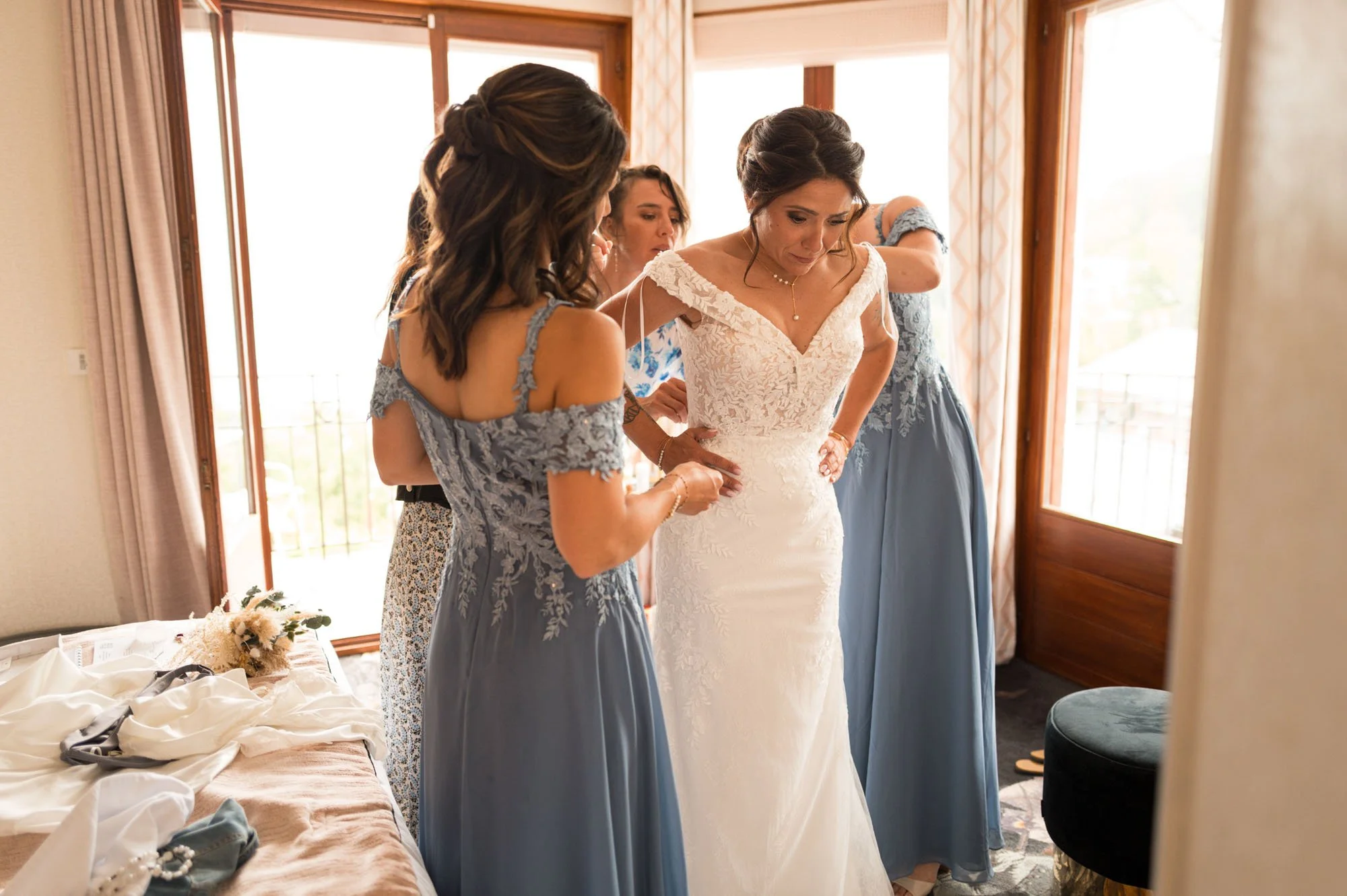 Une mariée en robe de mariage blanche avec deux femmes en robes bleues l'aident à ajuster sa robe. La scène se déroule dans une pièce lumineuse avec des fenêtres en arrière-plan.