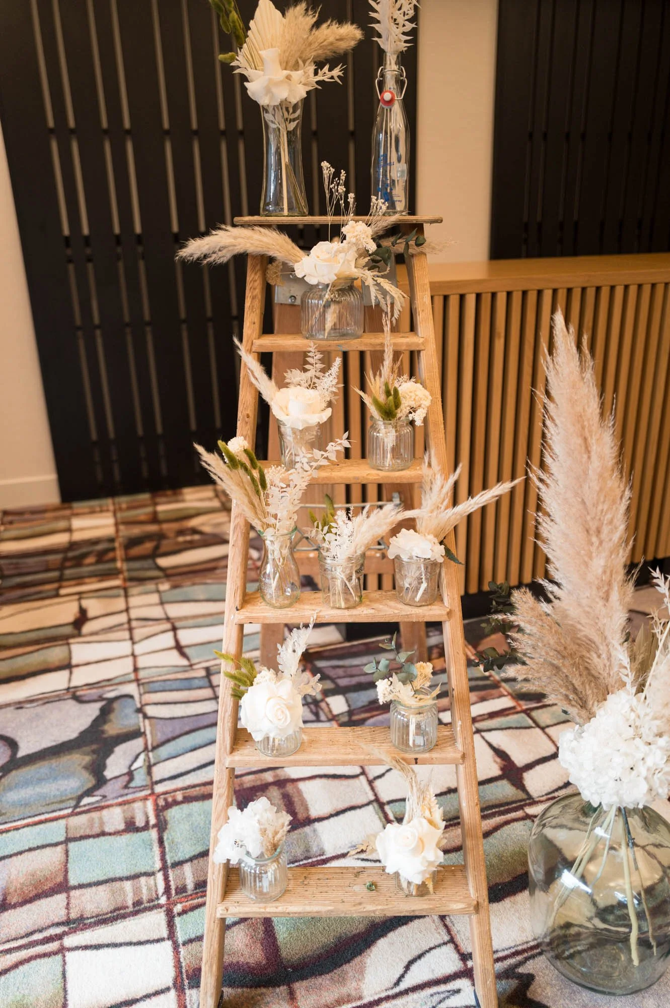 Échelle en bois décorée avec des vases en verre contenant des fleurs et des plantes séchées, avec un vase en verre volumineux à côté rempli de fleurs séchées.