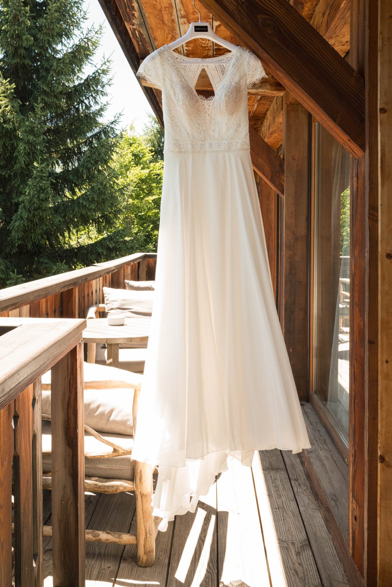 Une robe de mariée blanche suspendue à un porte-vêtements en plastique sur un balcon en bois, avec une vue d'arbres verts en arrière-plan, sous un toit en bois.