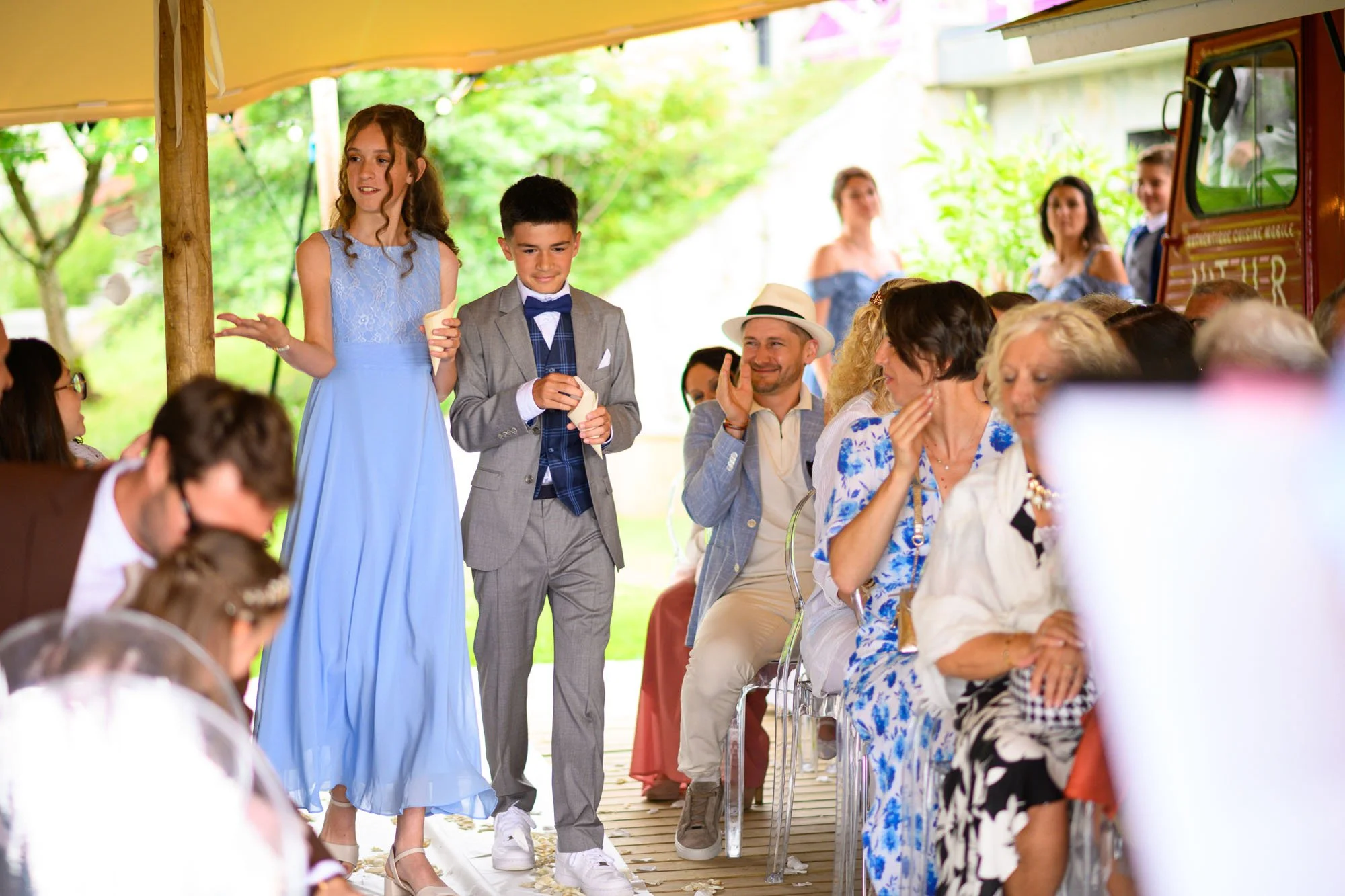 Un jeune couple habillé pour un mariage, la fille en robe bleue et le garçon en costume gris avec nœud papillon, marche parmi des invités assis sous un chapiteau lors d'une cérémonie en plein air.
