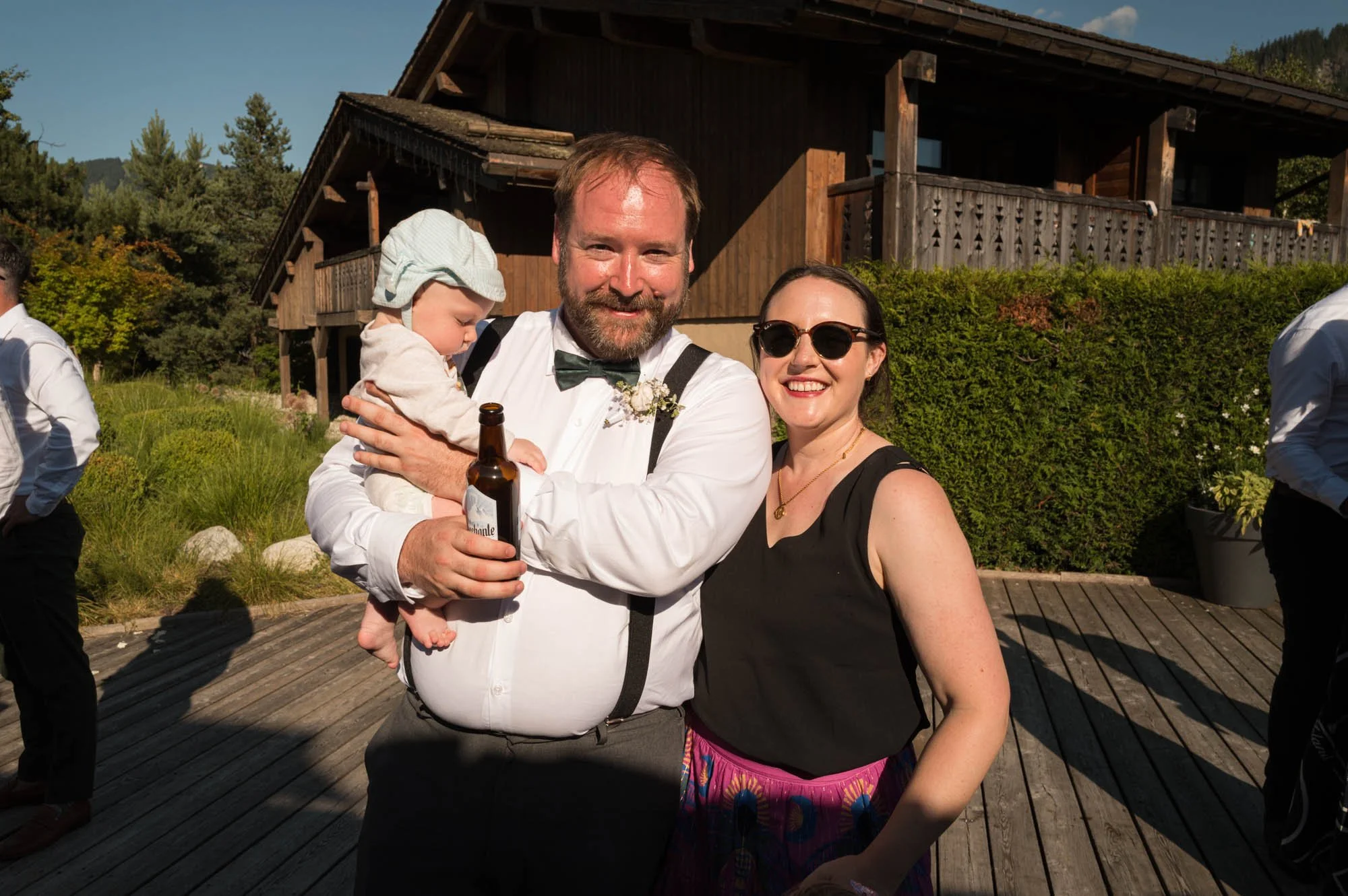 Un groupe de personnes lors d'une célébration en plein air, avec un homme tenant un bébé et une femme portant des lunettes de soleil, sous un ciel bleu et dans un cadre verdoyant.