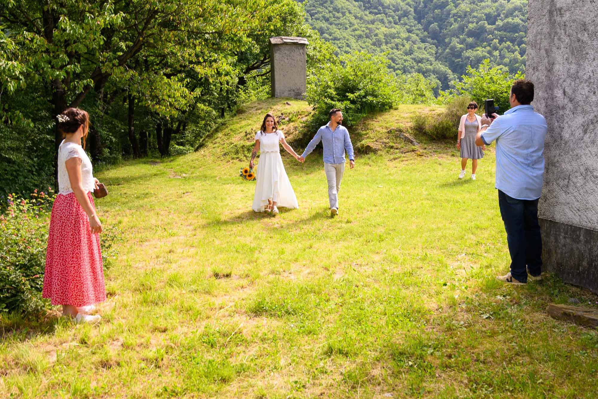 Un couple marié se promène main dans la main dans un champ vert, entouré d'arbres et de collines, avec trois autres femmes en arrière-plan, et un homme prenant une photo.
