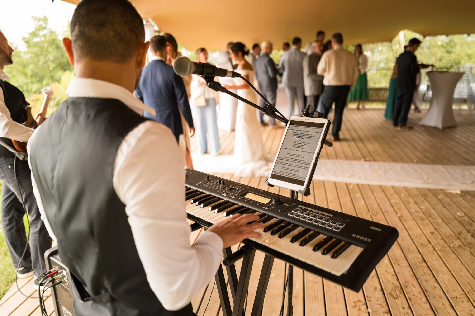 Musicien jouant du clavier lors d'une réception de mariage en plein air, avec un groupe de personnes en train de danser en arrière-plan.