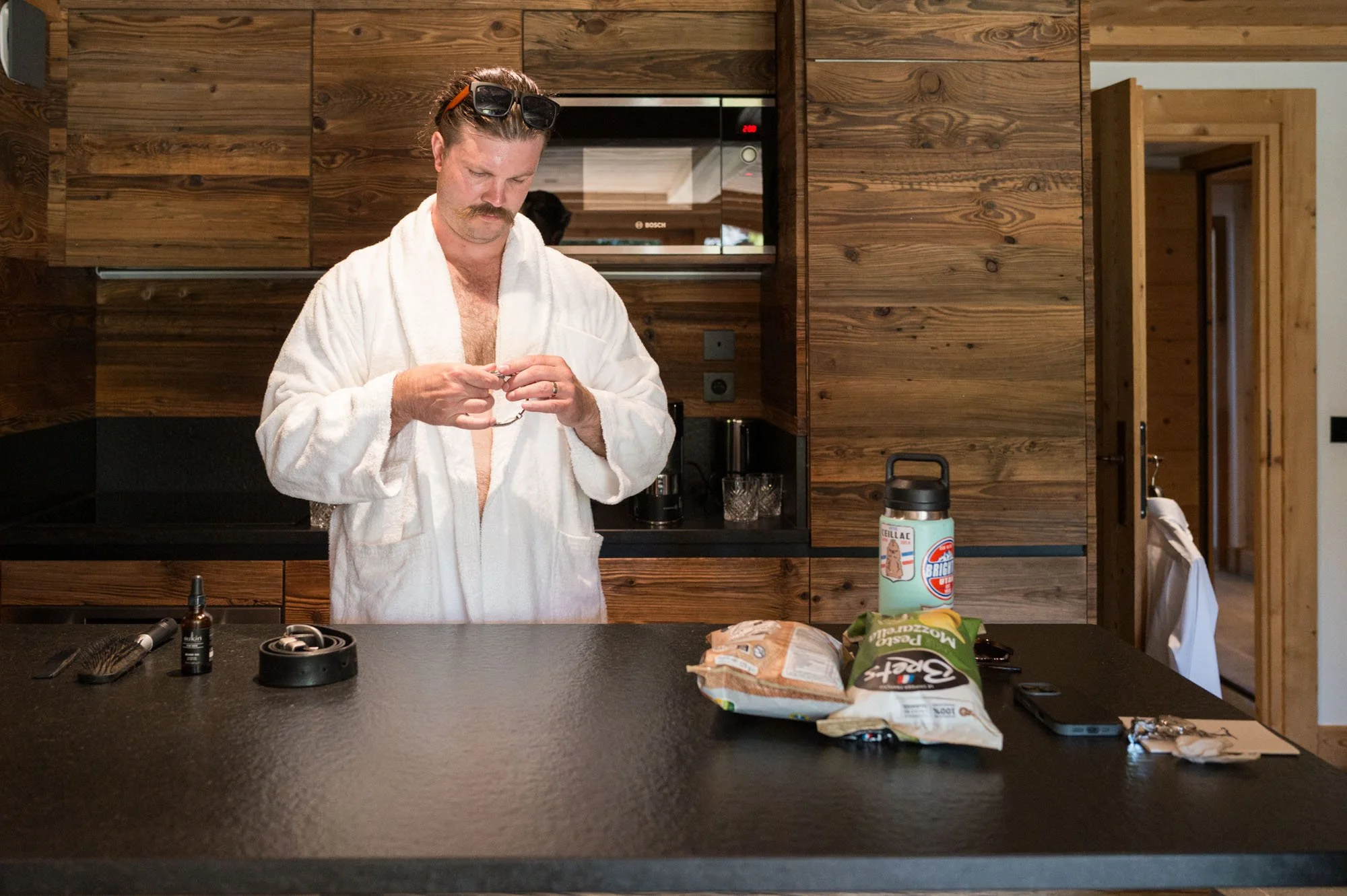 Un homme avec un peignoir blanc, des lunettes de soleil sur la tête, une moustache, qui regarde un petit objet dans ses mains dans une cuisine en bois.