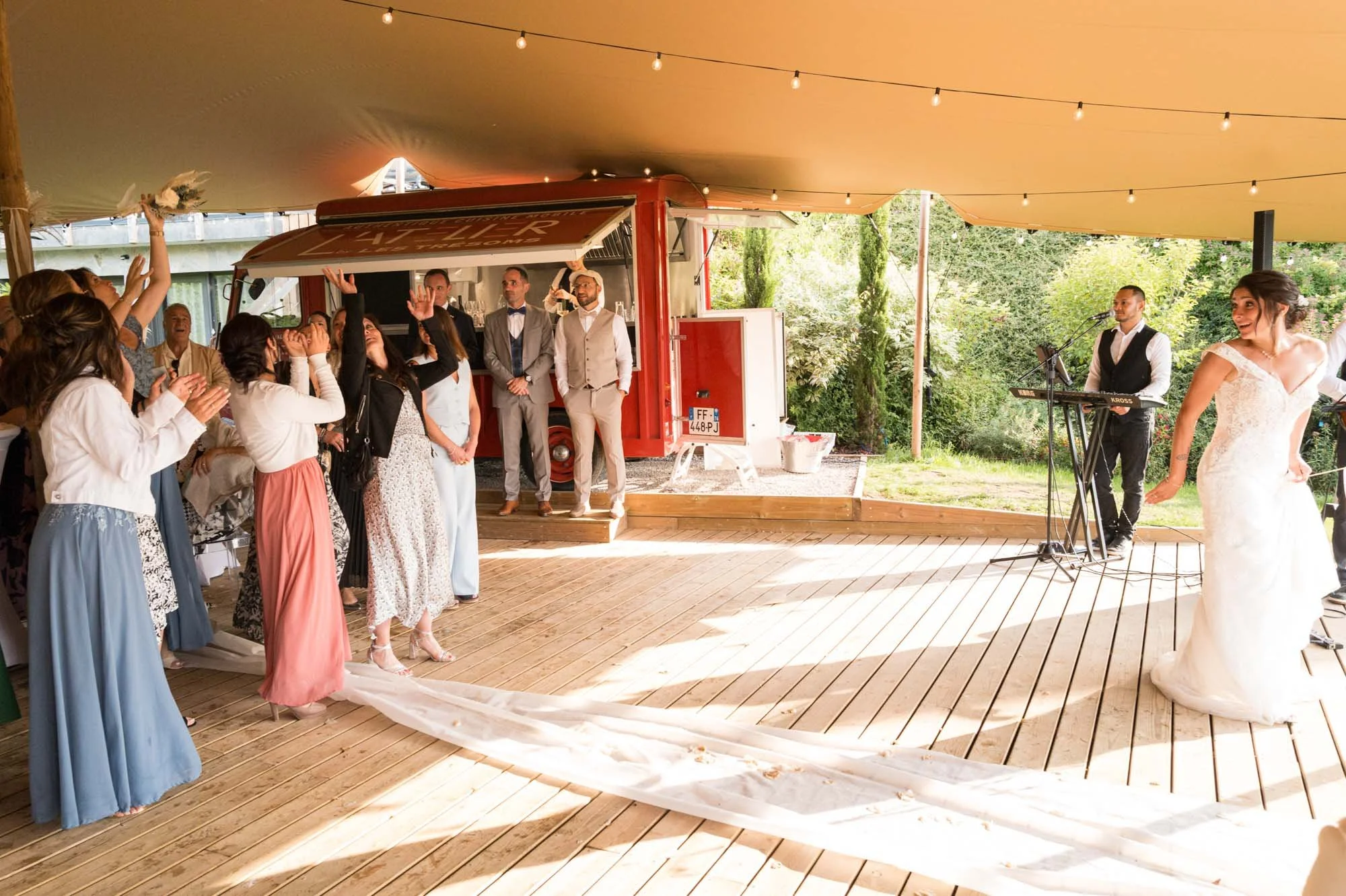 Un mariage avec la mariée en robe blanche, entourée d'invités et de musiciens dans une tente décorée de guirlandes lumineuses, avec un espace en plein air en arrière-plan.