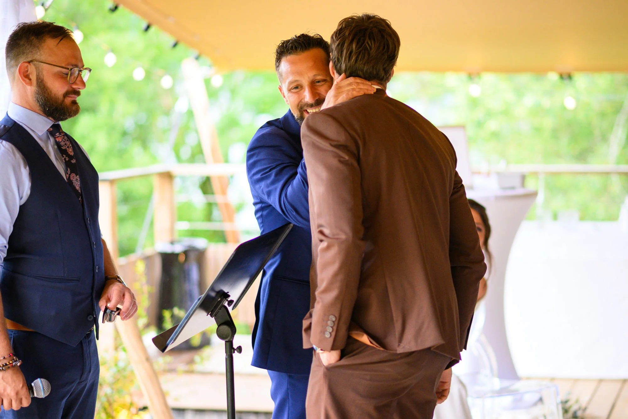 Deux hommes en costume bleu et marron s'embrassent lors d'une cérémonie en extérieur, tandis qu'un autre homme en veston bleu tient un micro à côté.