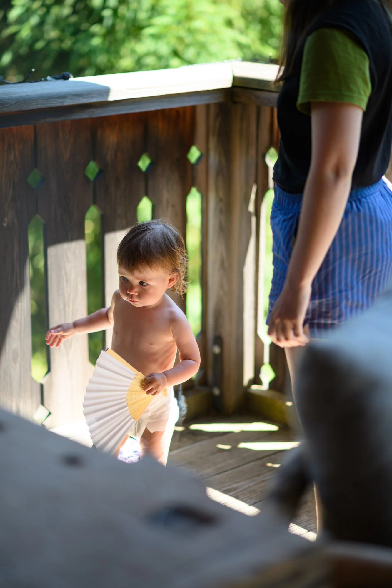 Un jeune enfant debout sur un pont en bois, tenant un éventail, avec une adulte à proximité dans un environnement extérieur verdoyant.